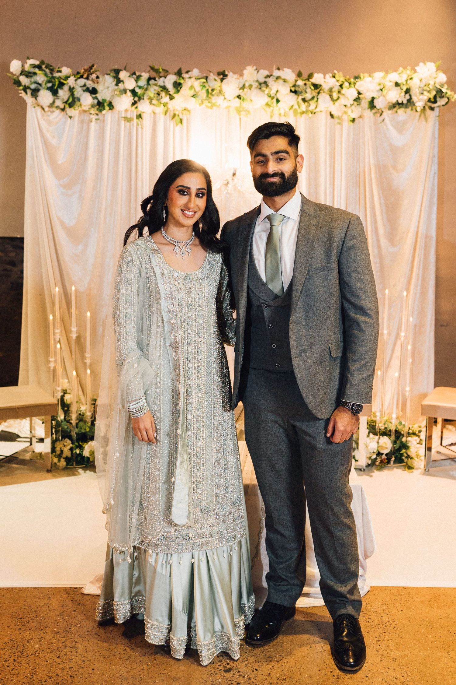 Couple poses at formal event with elegant floral backdrop and white curtains in formal attire.