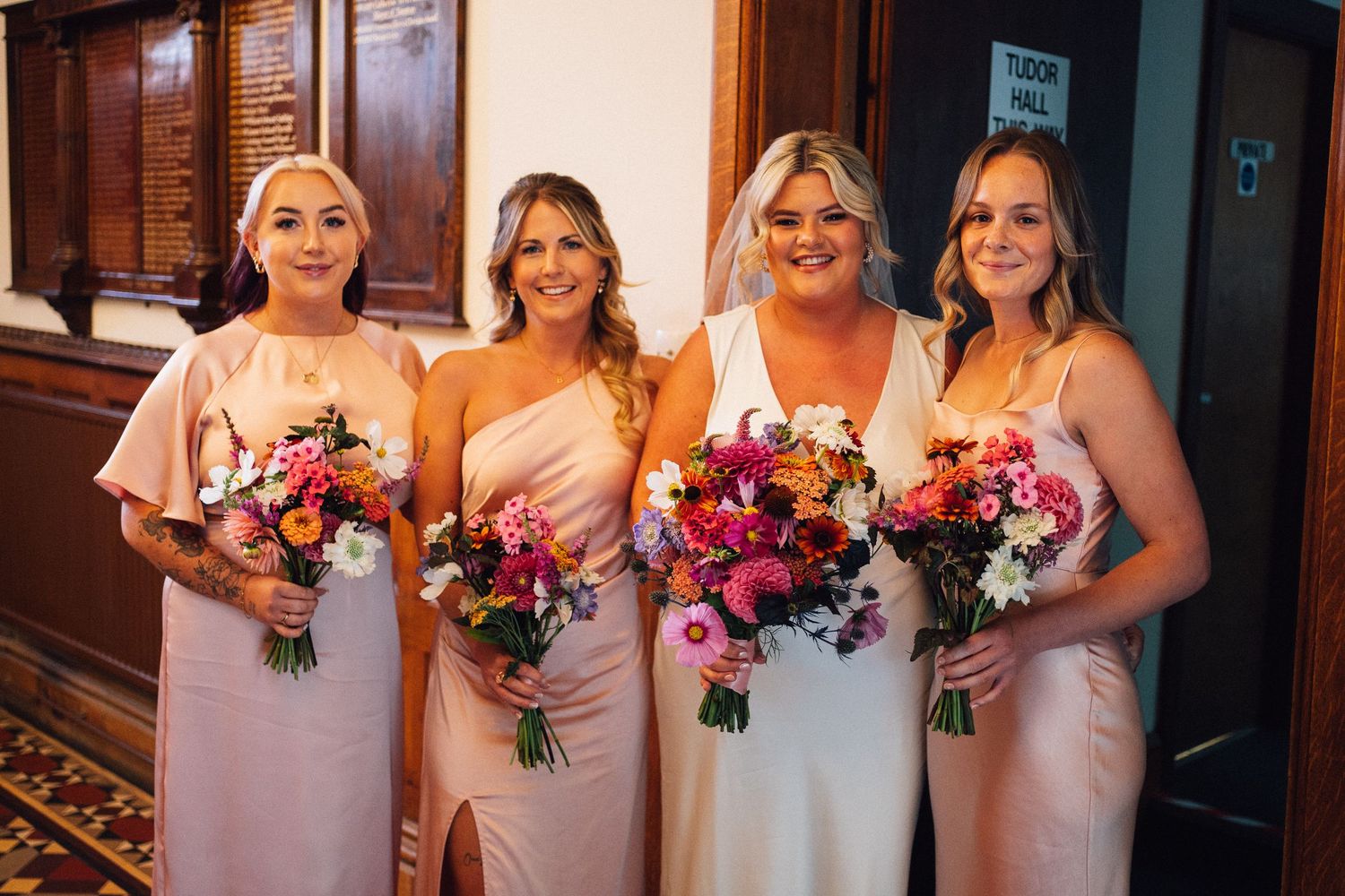 Bridesmaids in blush pink dresses hold vibrant pink and purple bouquets while posing together at a wedding.