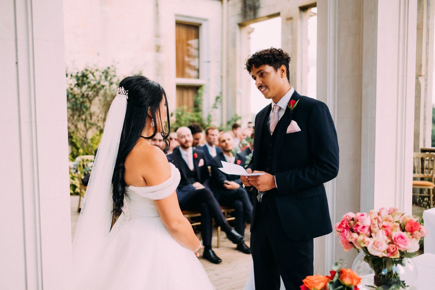 A bride and groom exchange vows during an elegant outdoor wedding ceremony surrounded by white columns and pink flowers.