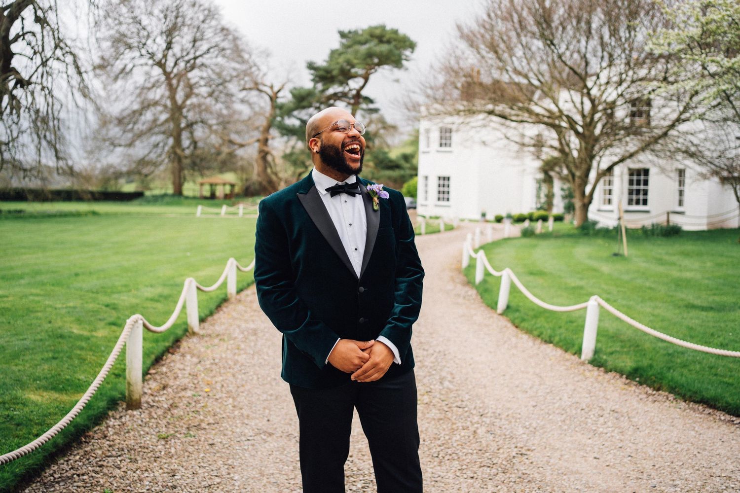 A person in a black tuxedo and bow tie stands on a gravel path with a white estate house and green lawn in the background.
