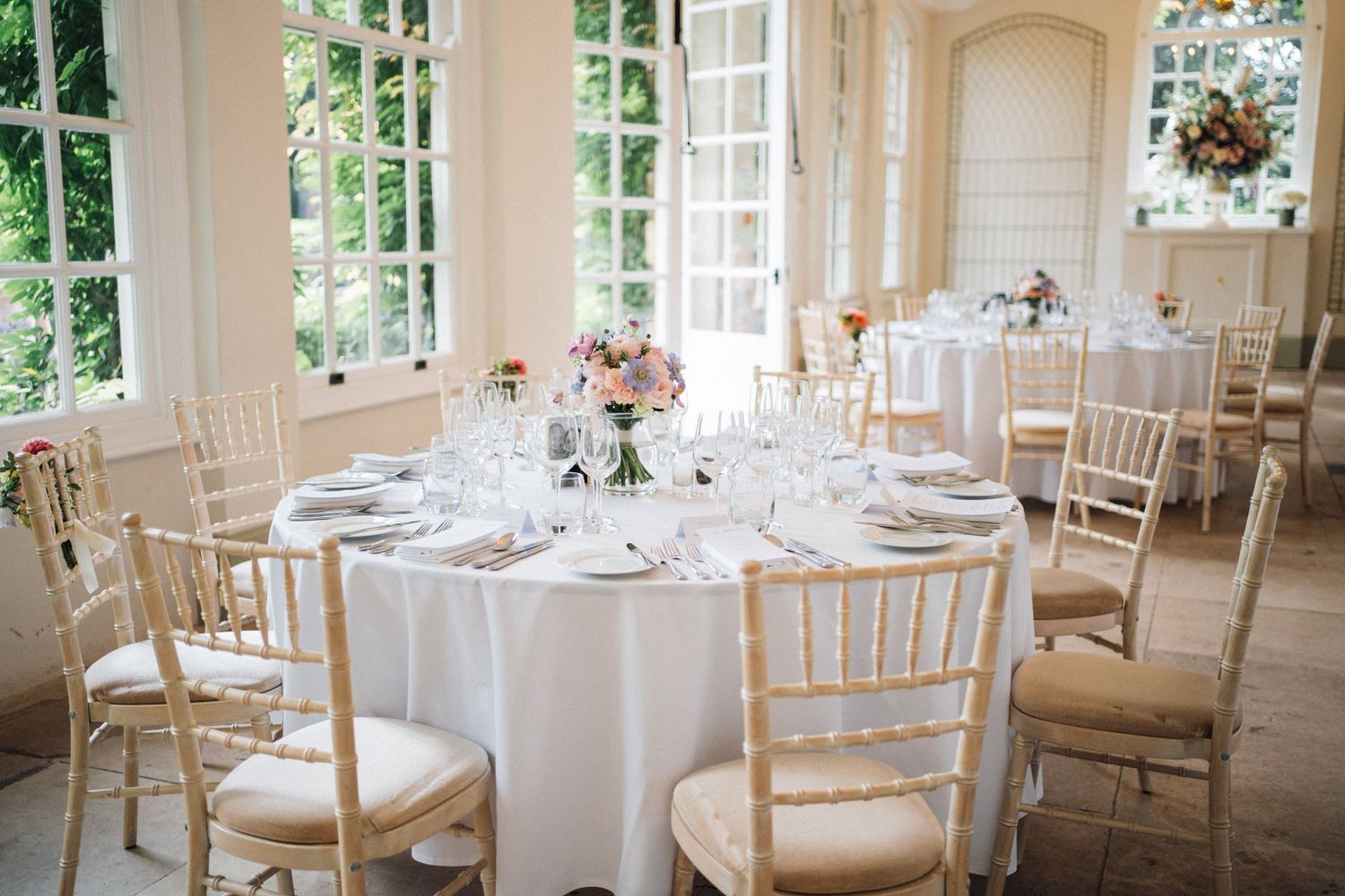 Elegant wedding reception room with round tables, white linens, gold Chiavari chairs, and floral centerpieces in a bright venue.