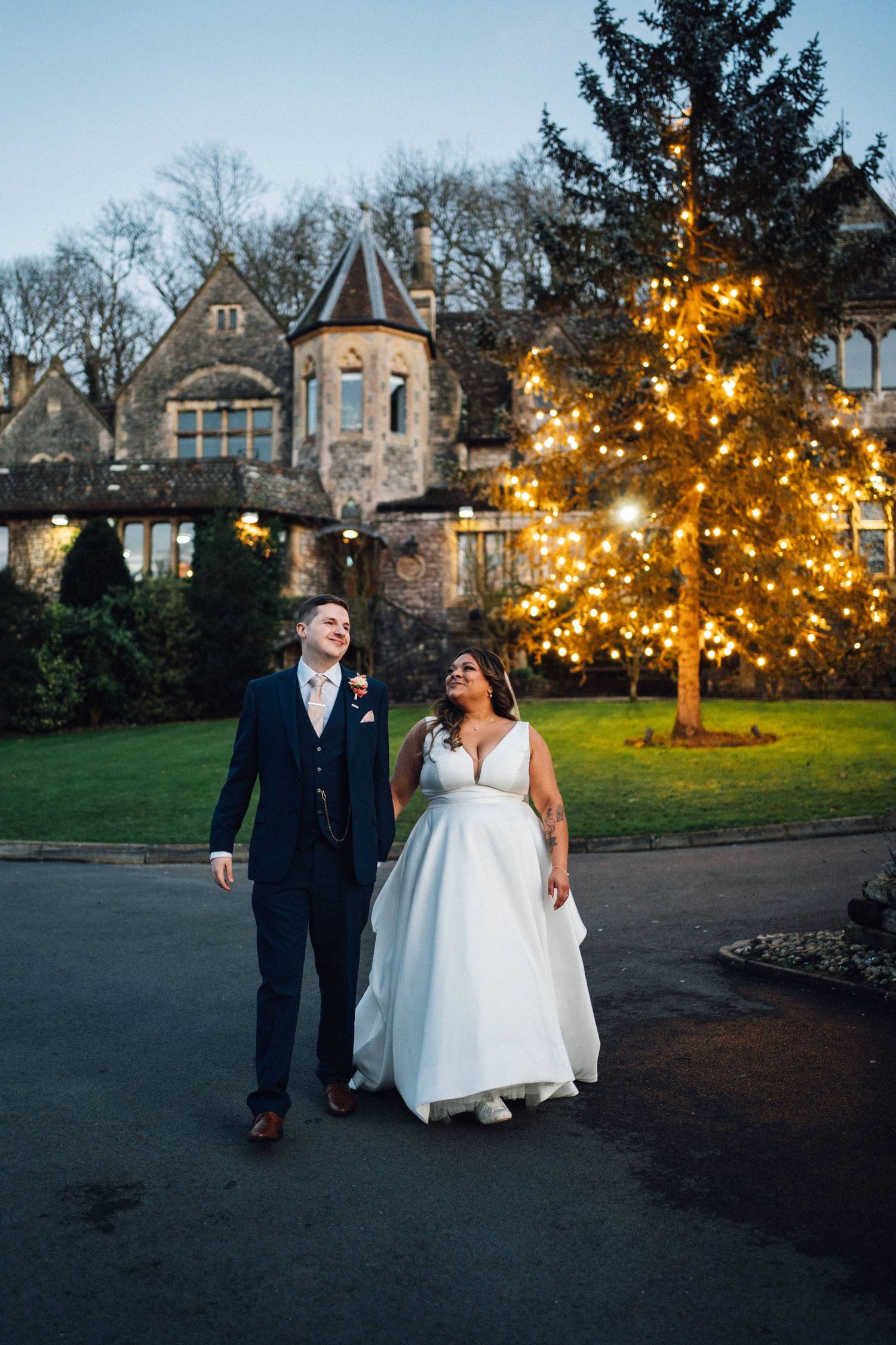 A couple walks along a path at dusk with glowing trees and historic buildings creating a romantic backdrop.