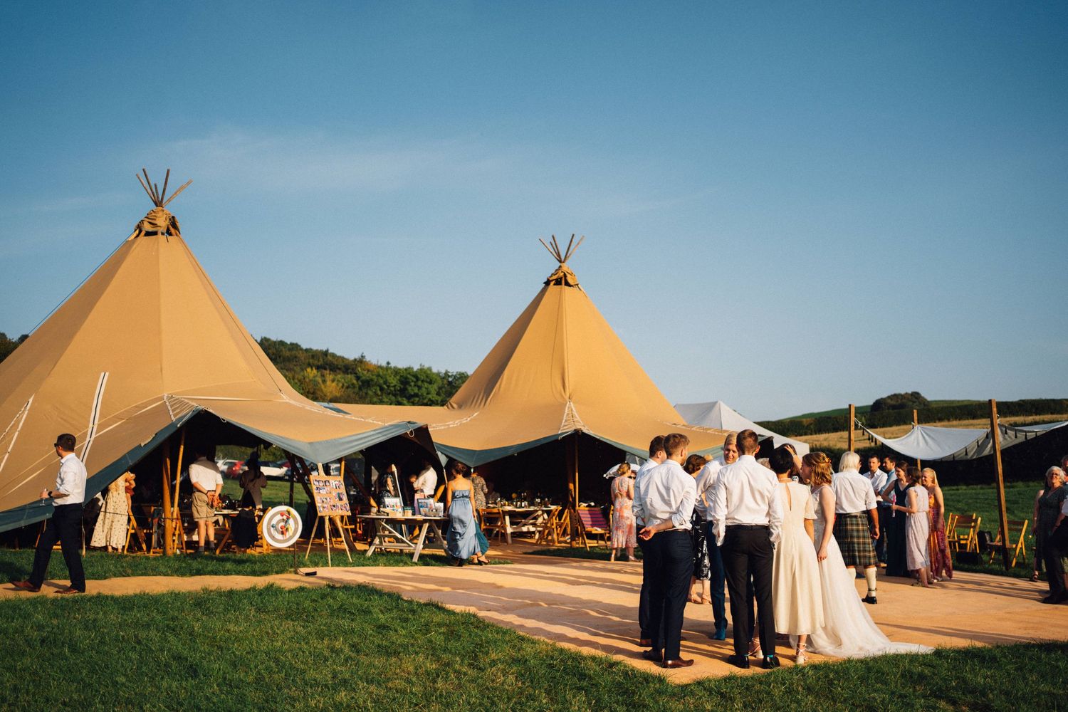 Outdoor wedding reception with guests gathered near large tipi tents on a sunny day in a scenic countryside setting.
