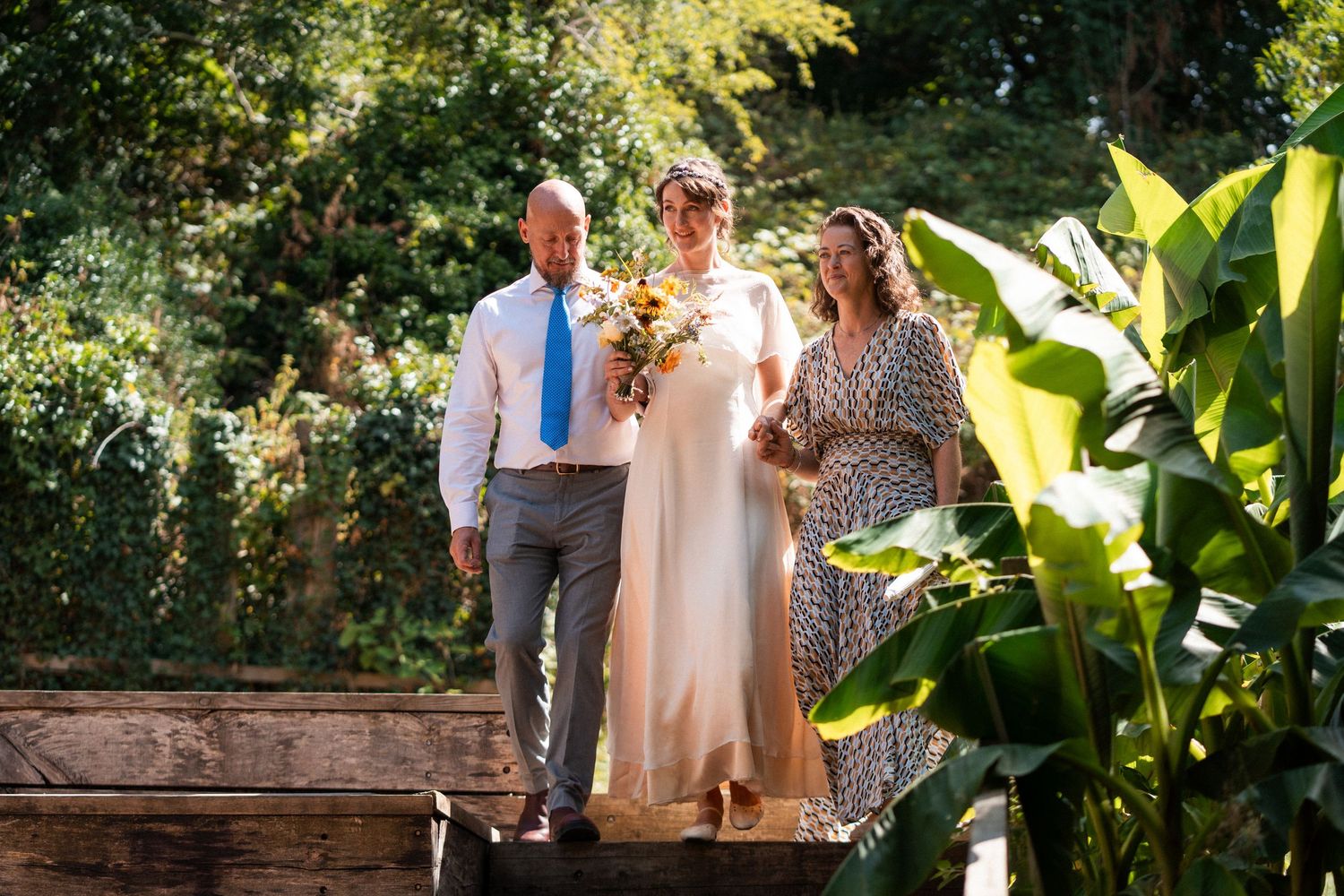 Bride in flowing gown holds bouquet flanked by groom and guest on wooden steps surrounded by lush tropical greenery.