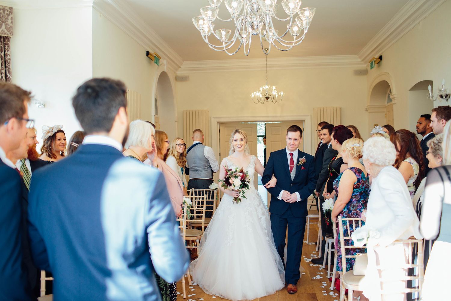 Wedding ceremony guests gather in an elegant room with chandeliers and natural light streaming through windows.