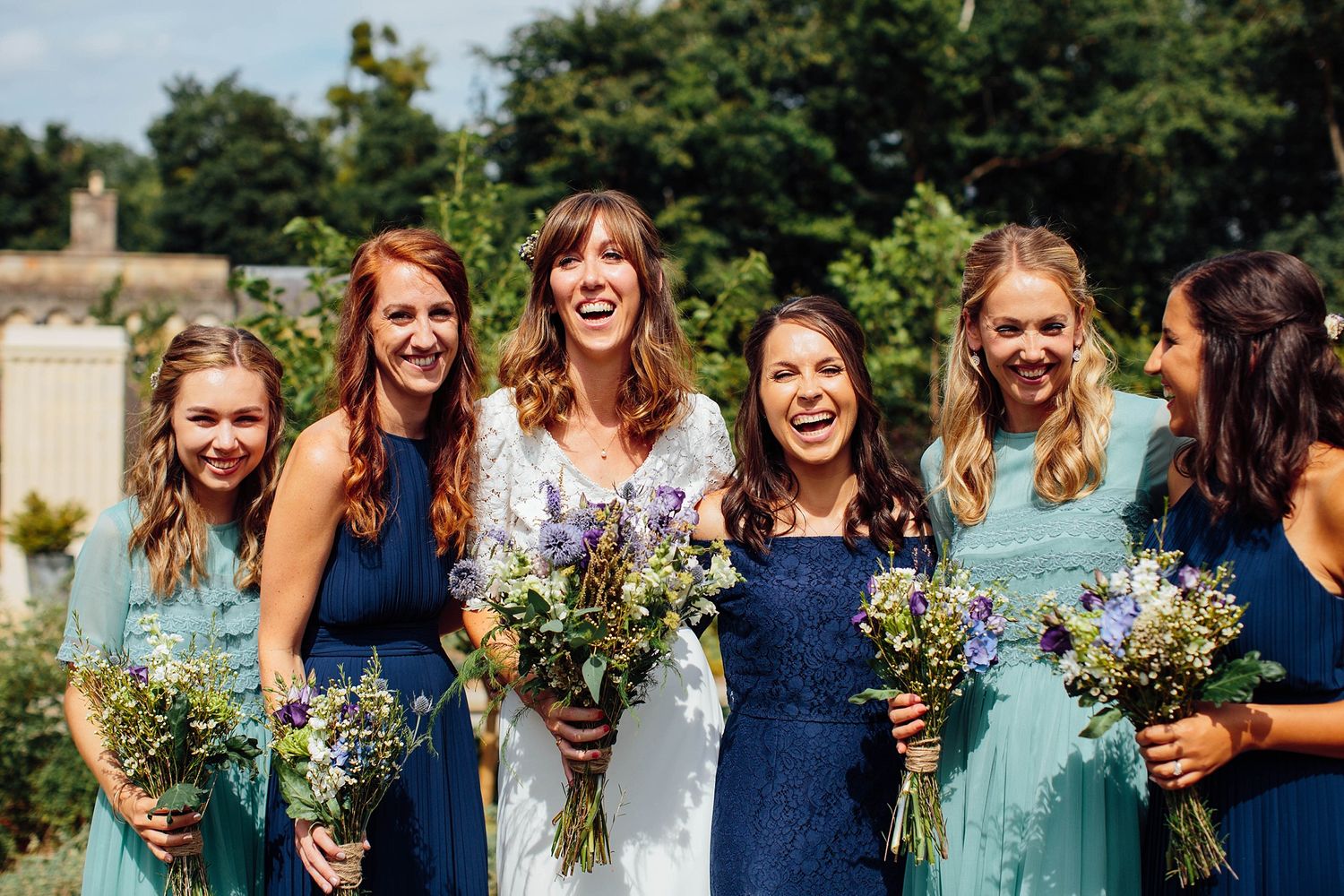 A joyful bridal party in blue and mint dresses holds wildflower bouquets in a garden.