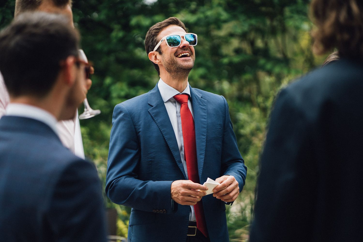 Groomsmen in dark suits and red ties chatting during outdoor wedding reception.