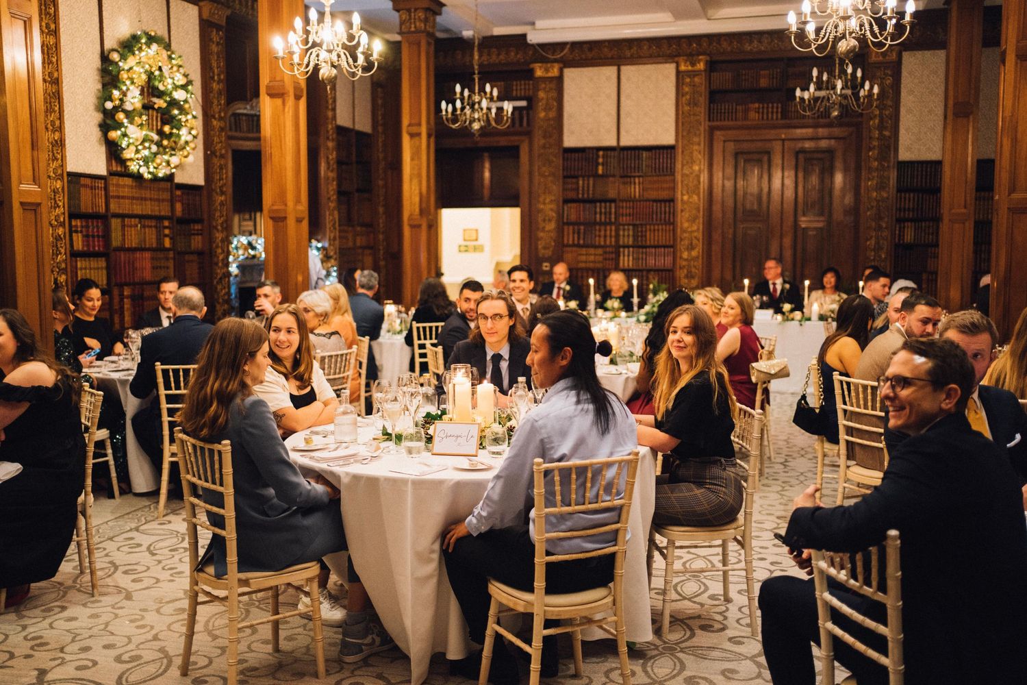 Wedding reception panorama showing guests dining under crystal chandeliers in a grand hall.