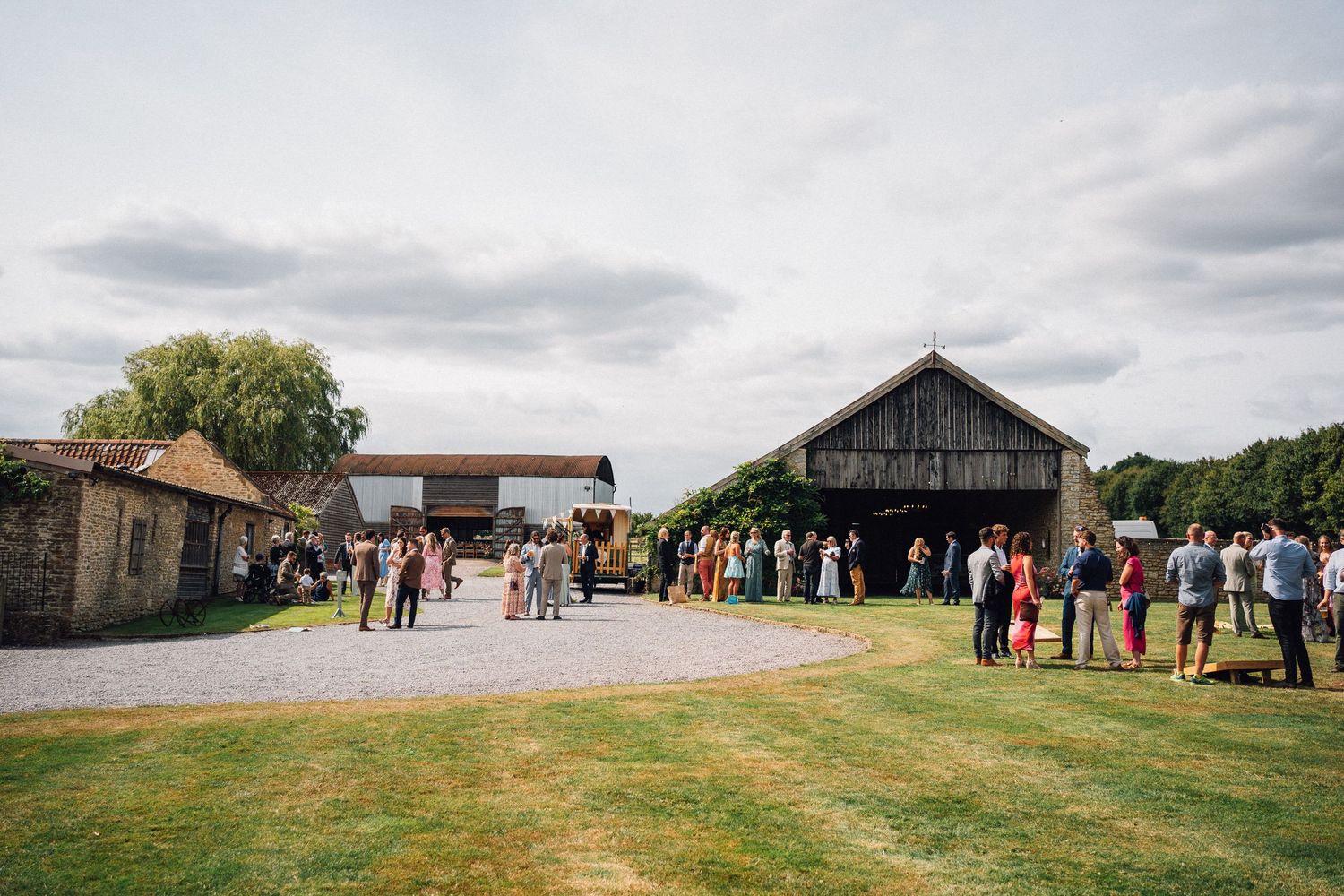 Panoramic view of wedding guests gathering outside a rustic barn venue on a cloudy day.