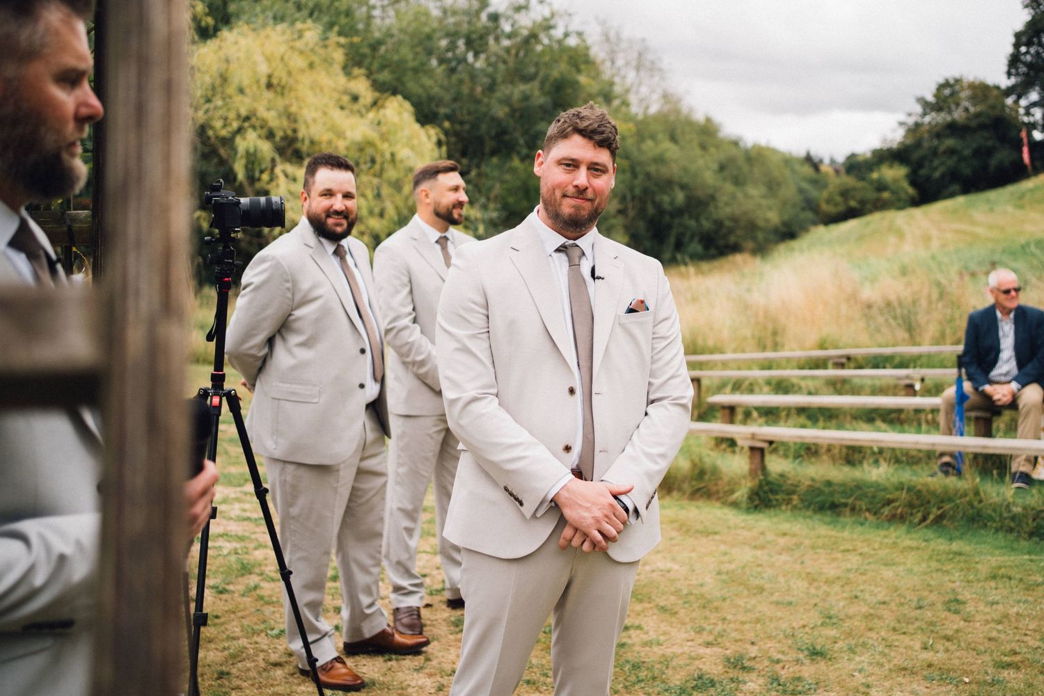 Groomsmen in light beige suits gather before the ceremony in an outdoor setting.