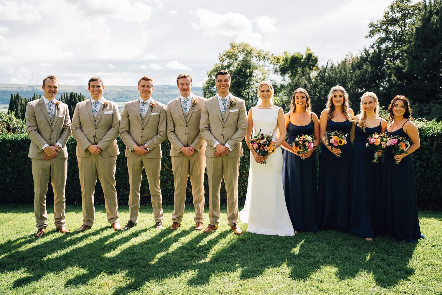 Wedding party lines up for formal group photo with bridesmaids in navy and groomsmen in tan.