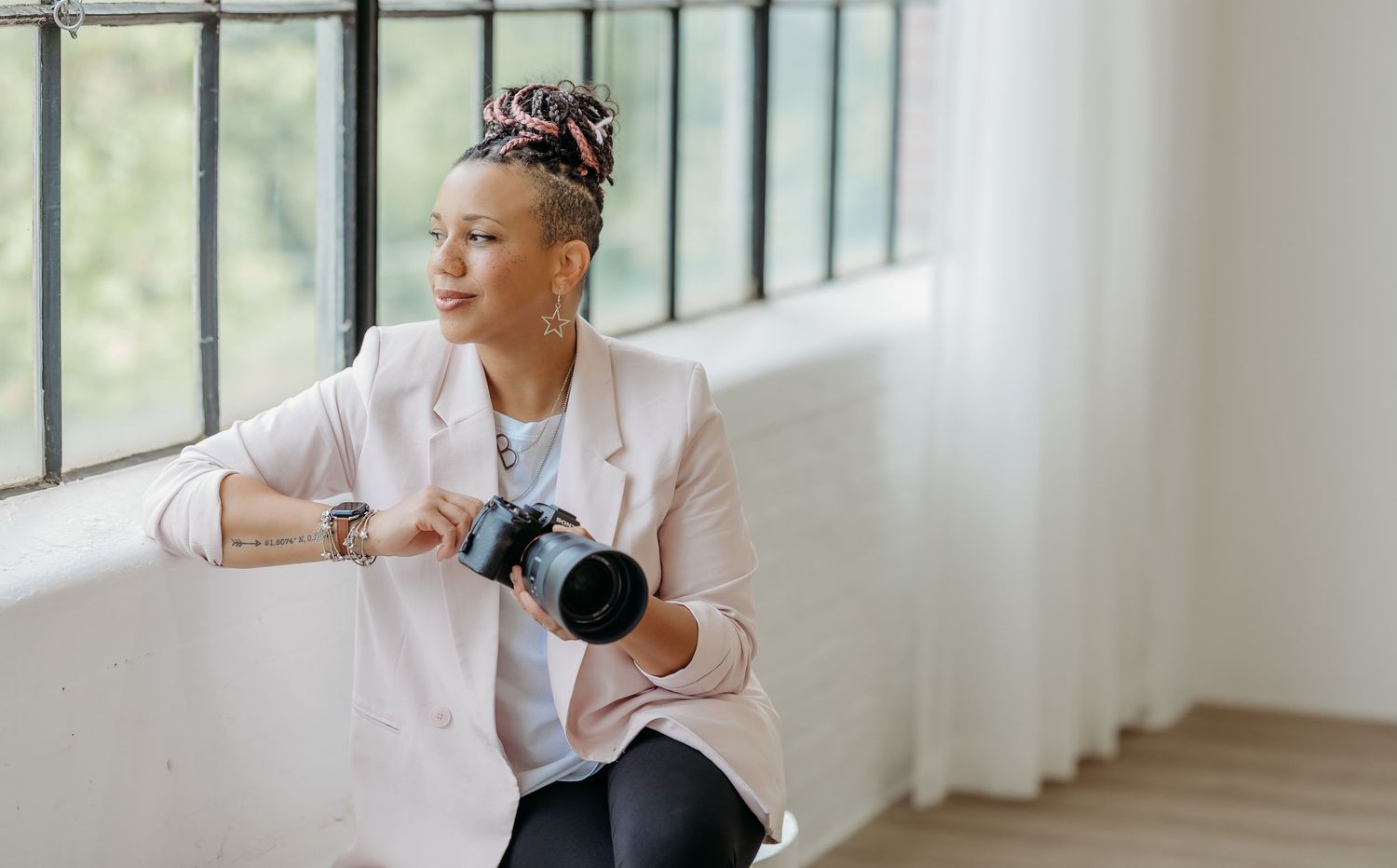 Professional photographer in pink blazer sits by window with camera in minimalist bright studio space.