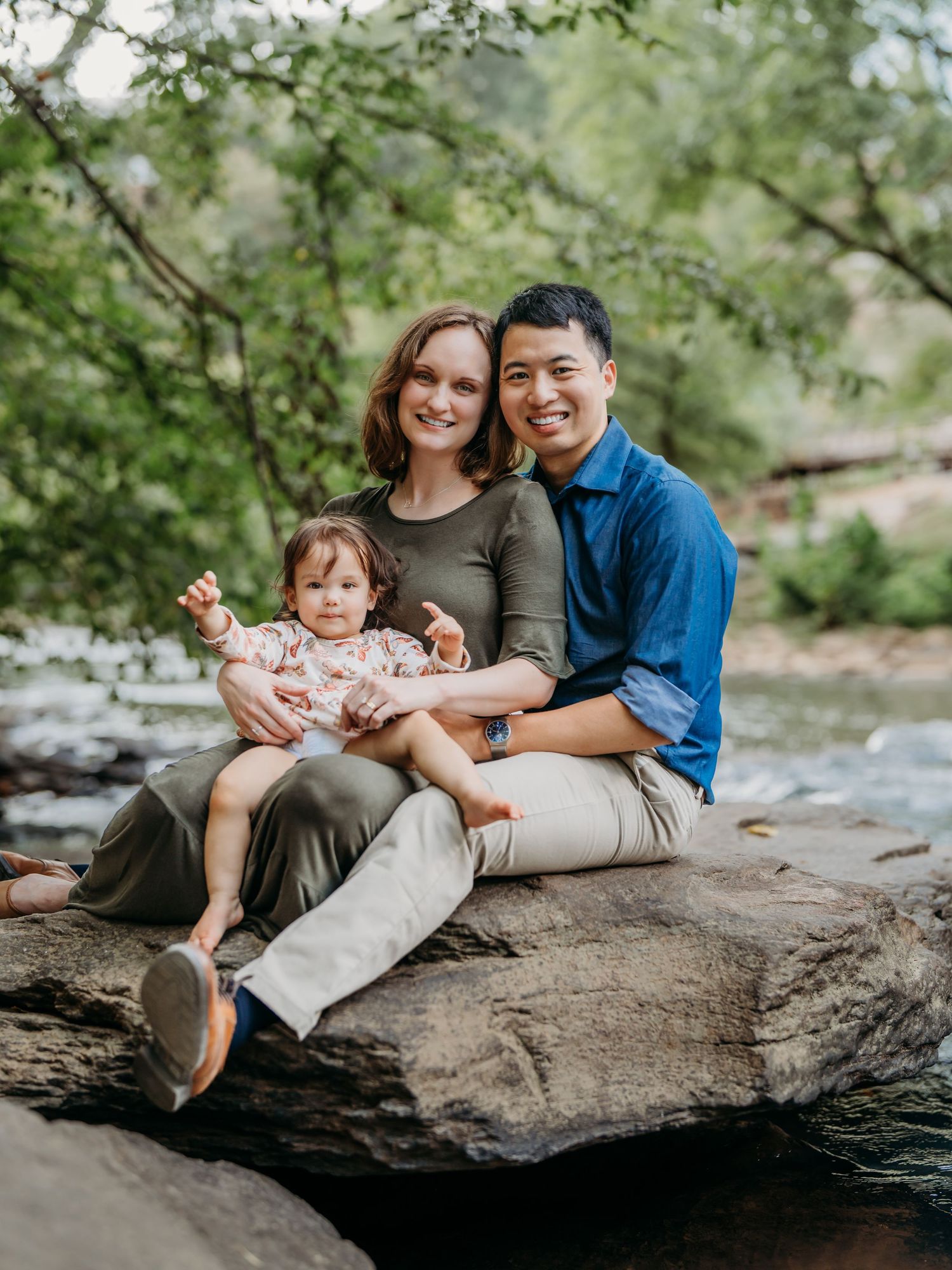 Family wearing casual clothing sits together on large rock formation by stream in green natural setting.
