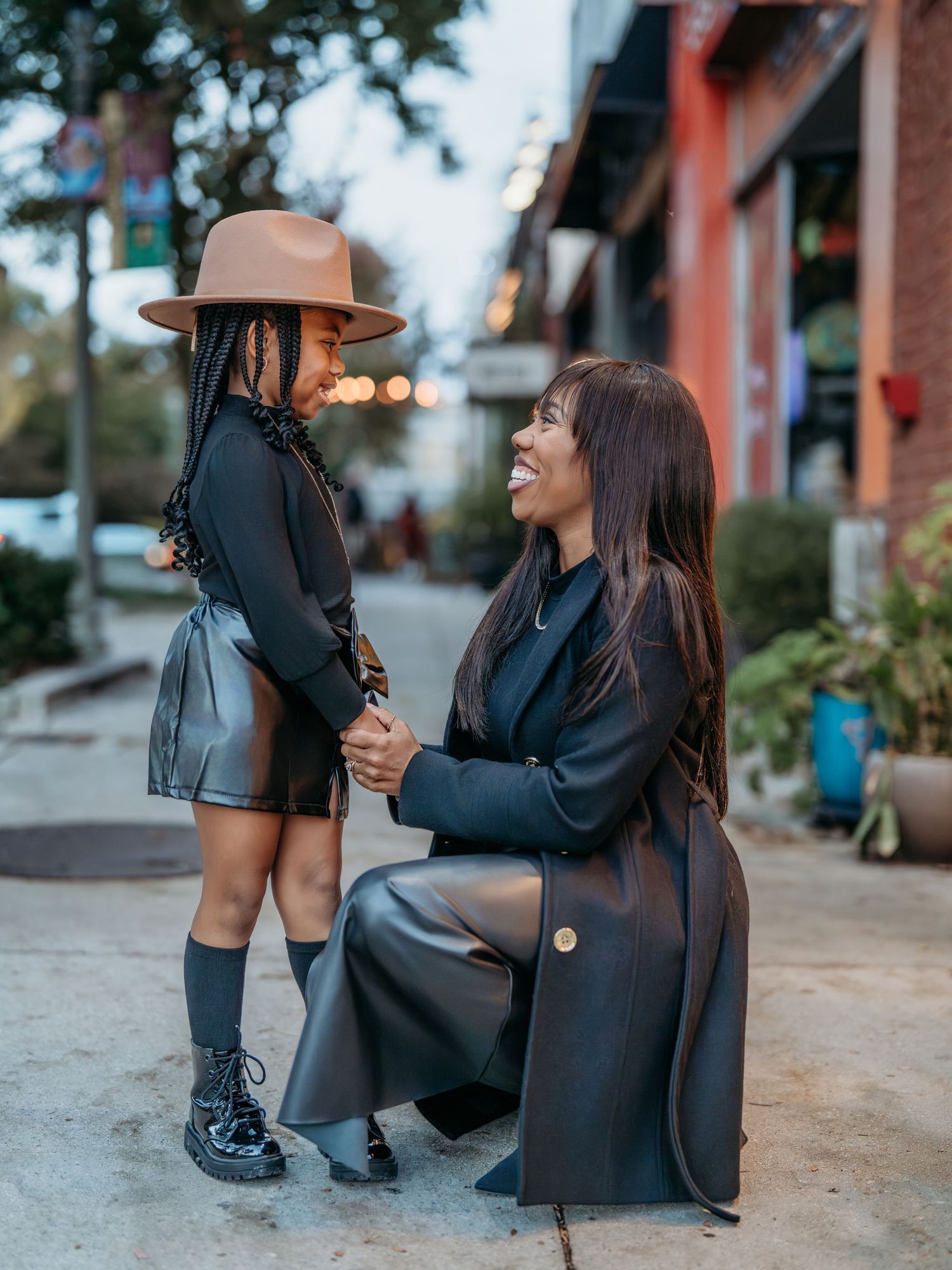 A heartwarming moment of a mother and daughter captured on a city sidewalk with matching black outfits, a brown hat, and autumn decor.