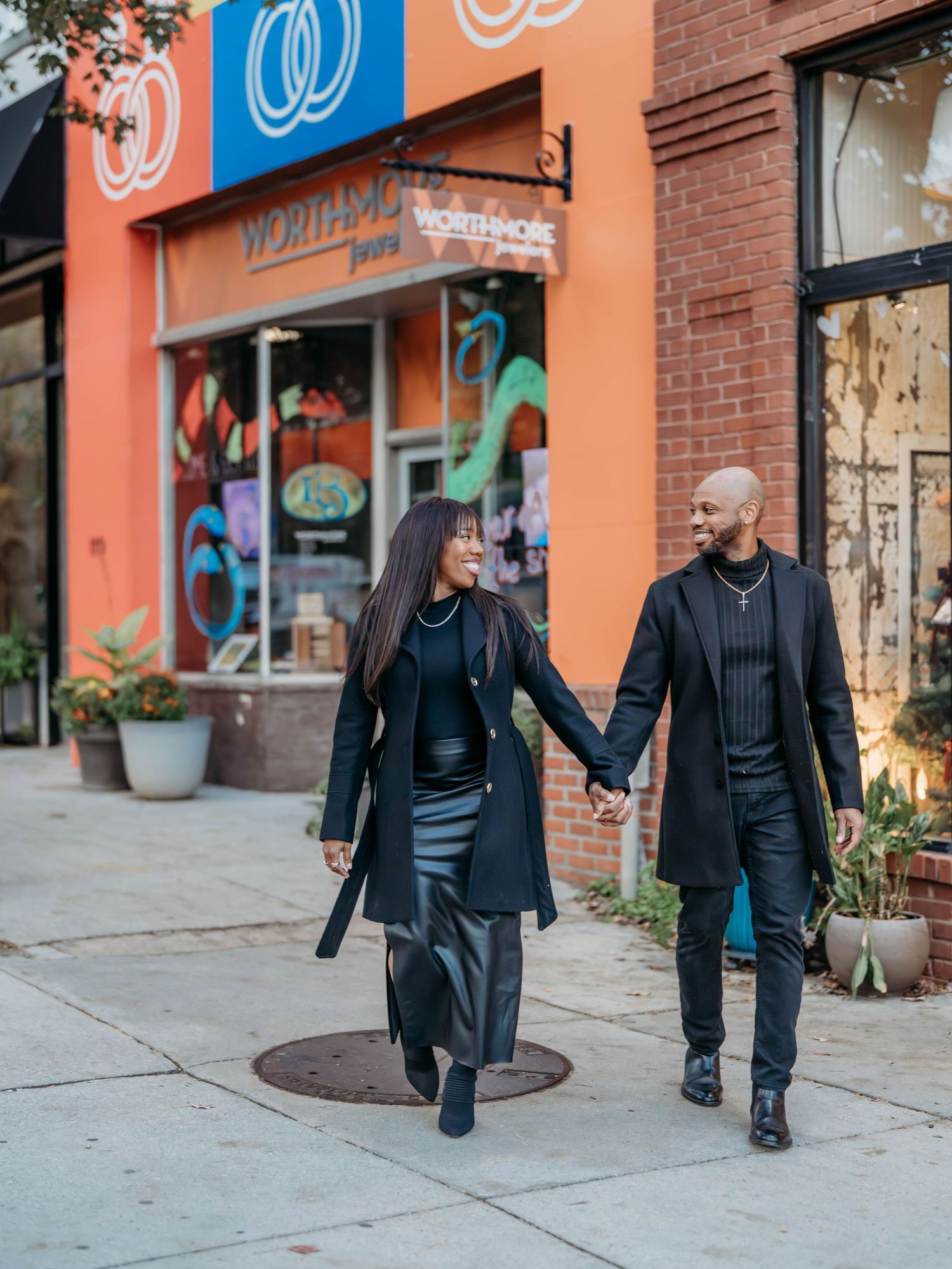 Two people in dark outfits walk past a brick storefront with blue and orange signage in an urban setting.