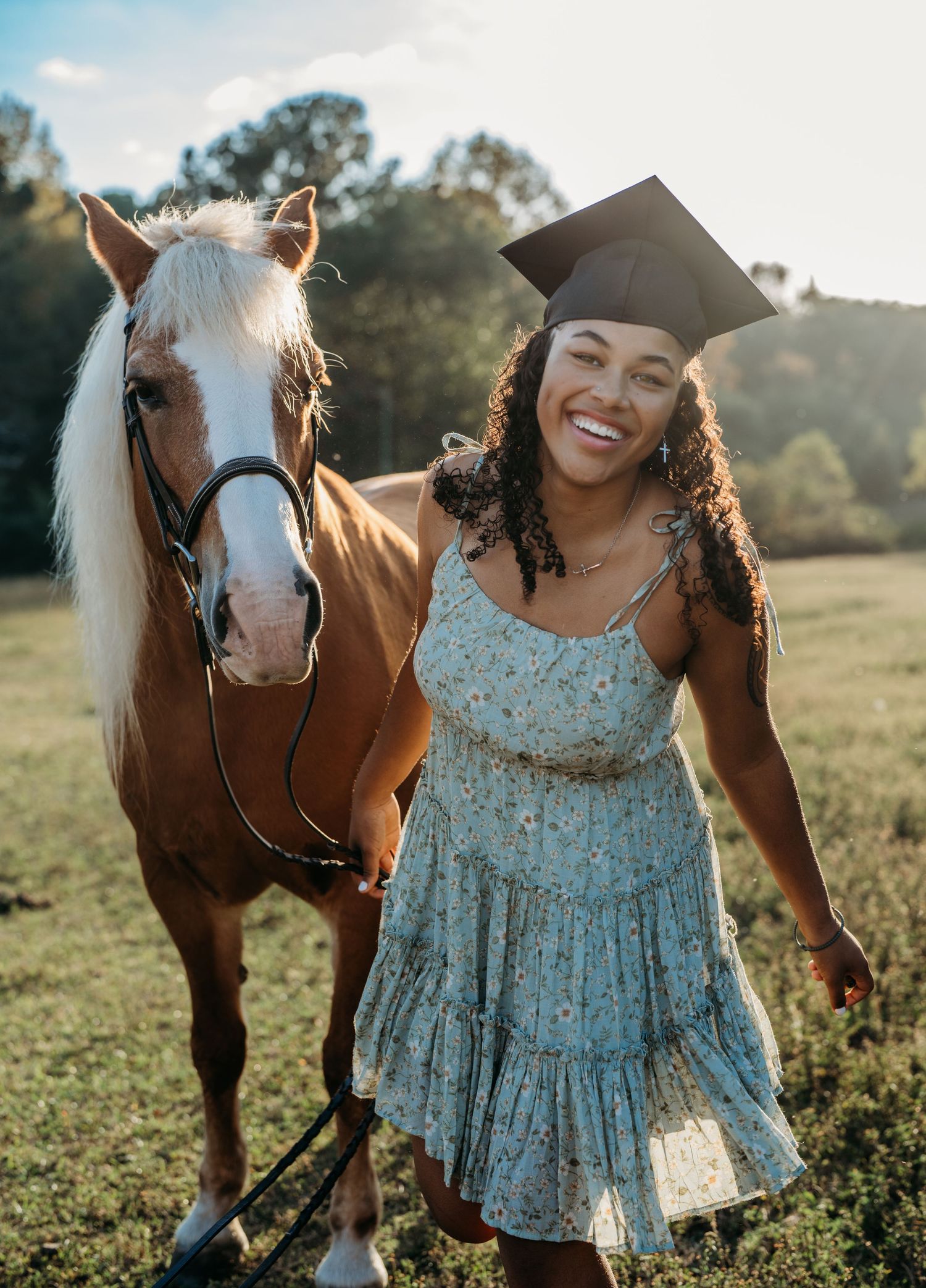 A brown and white horse stands beside someone wearing a floral blue dress in a sunlit meadow during golden hour.