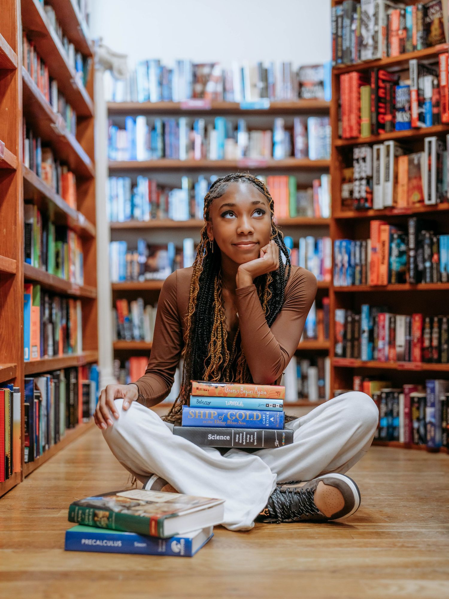 Young person with braids lying on library floor surrounded by wooden bookshelves while reading and relaxing.