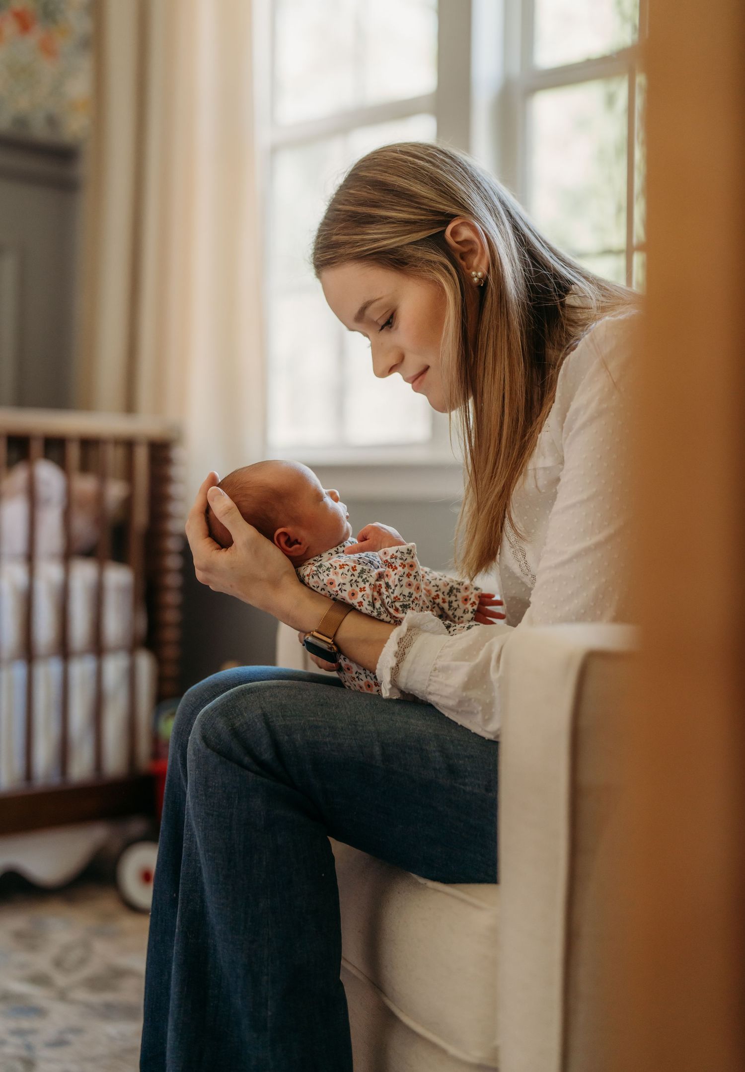 A mother holds and kisses her sleeping baby in a nursery setting near a window with soft natural light.