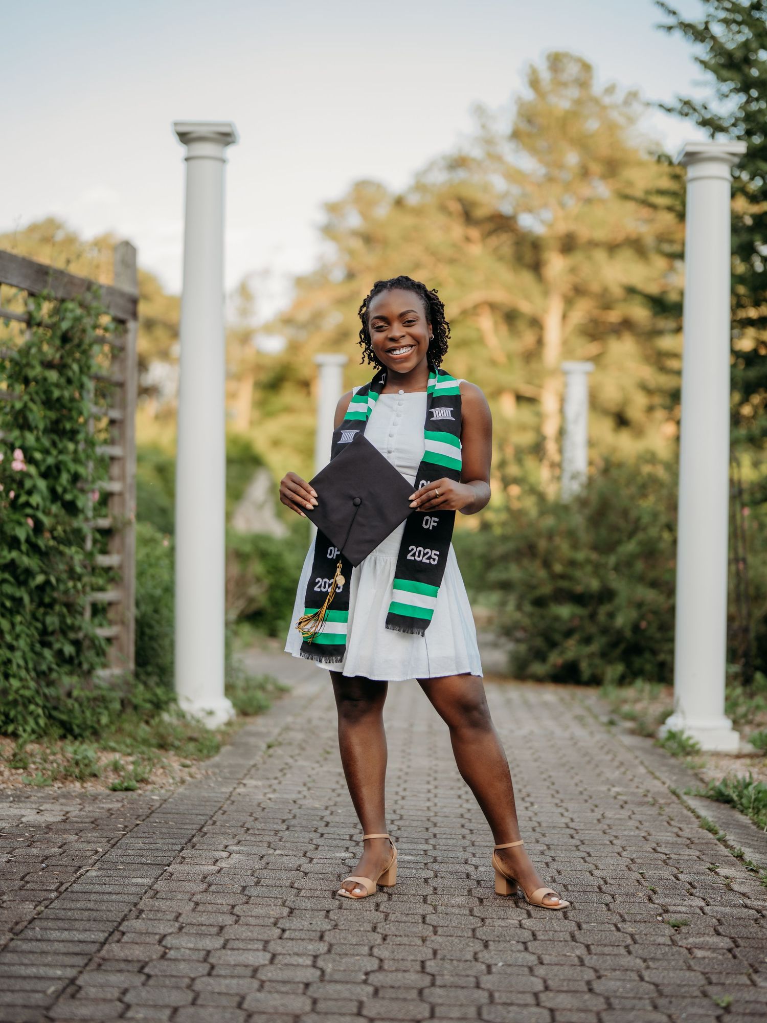 A graduate in a white dress and graduation stole poses confidently on a brick pathway lined with white columns.