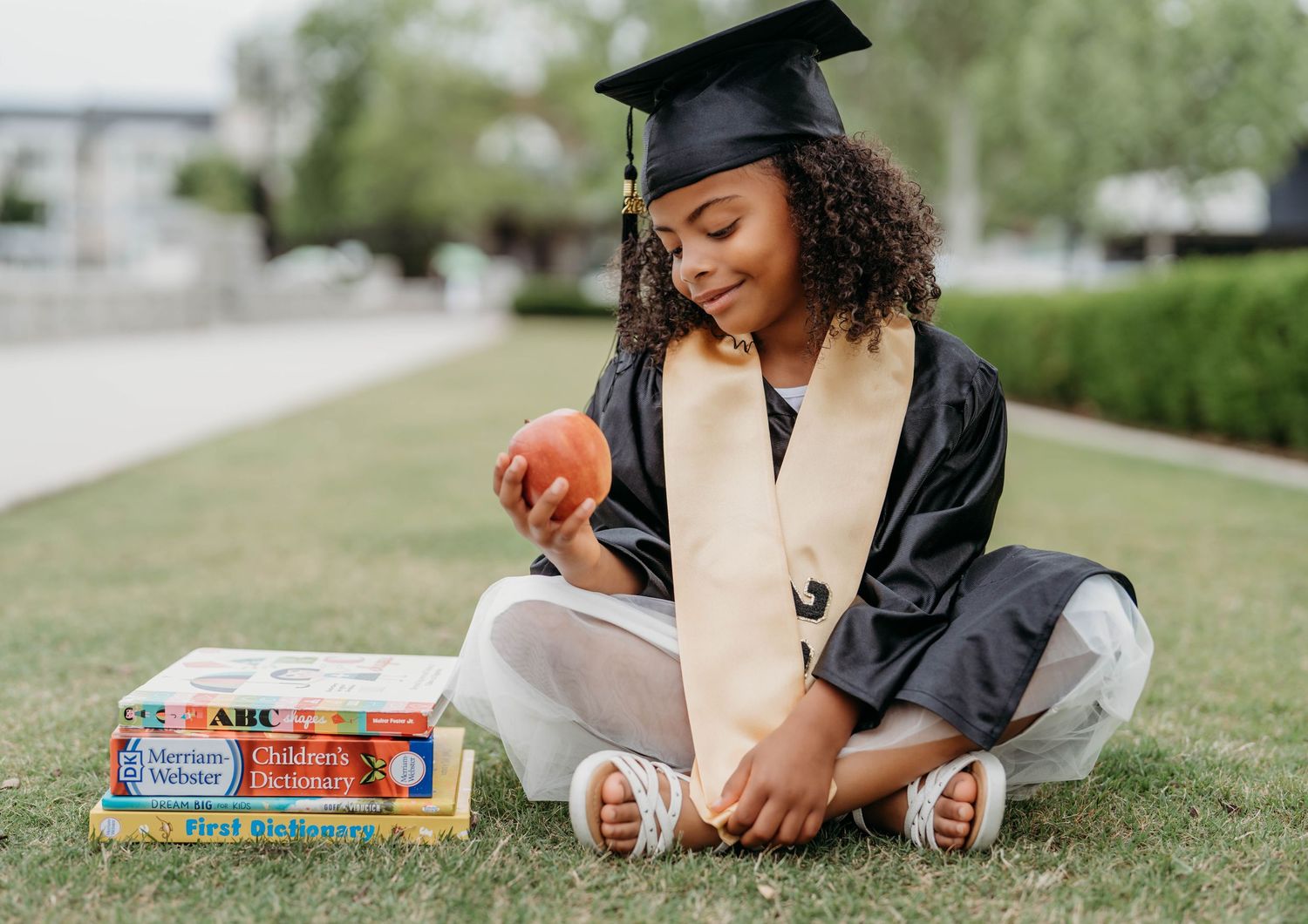 Young girl in graduation cap and gown sitting on grass holding an apple with childrens books beside her.