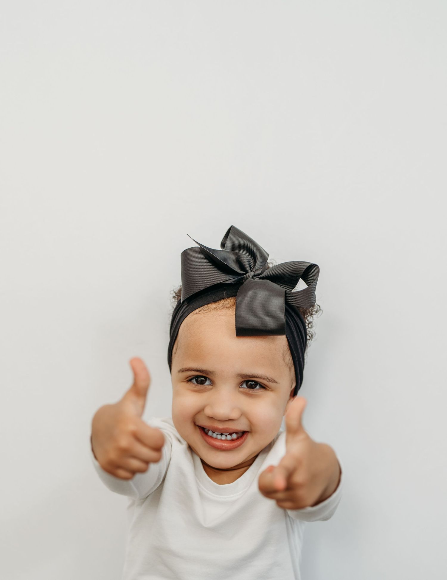 A child wearing a black bow and white shirt shows thumbs up signs while smiling at the camera.