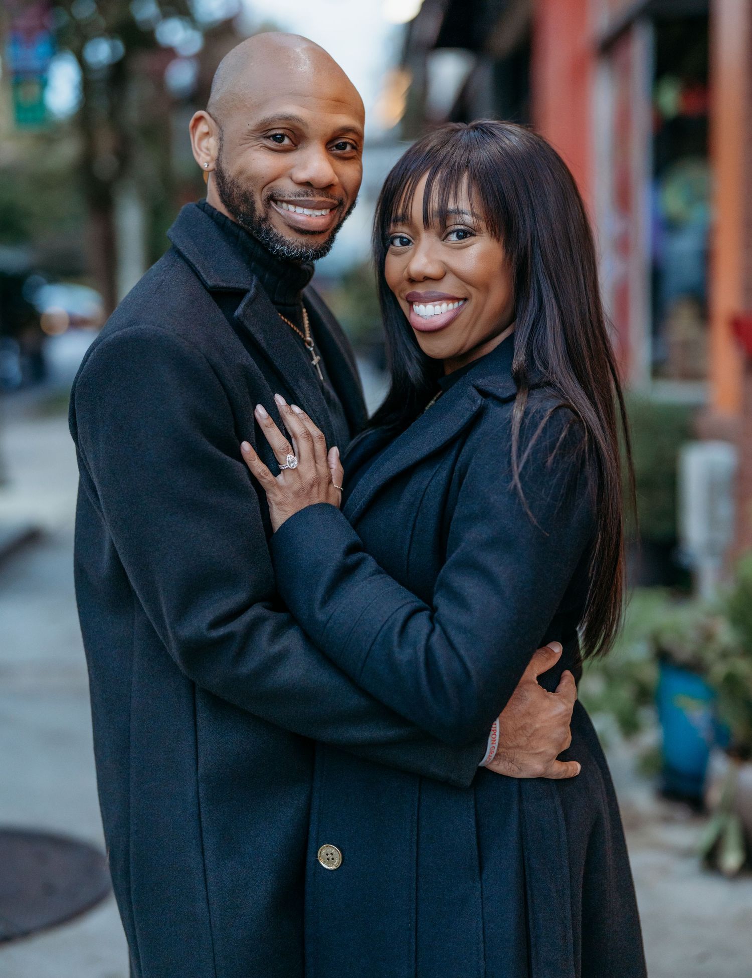 A couple shares a tender embrace while wearing black coats outdoors during an engagement photoshoot.