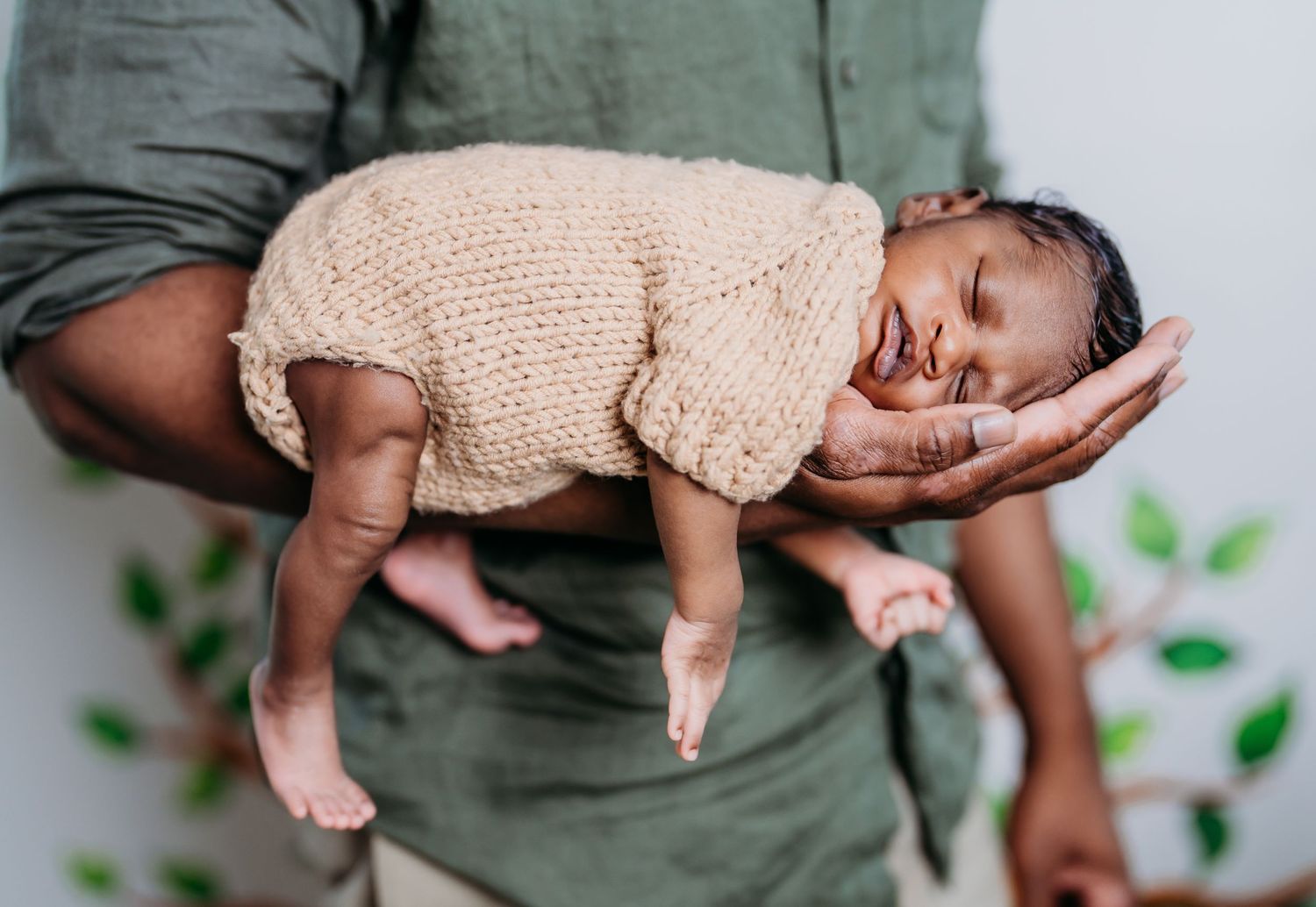A newborn baby wearing a peach knit outfit lays sleepily on a dark cushion as green plants peek into the frame.