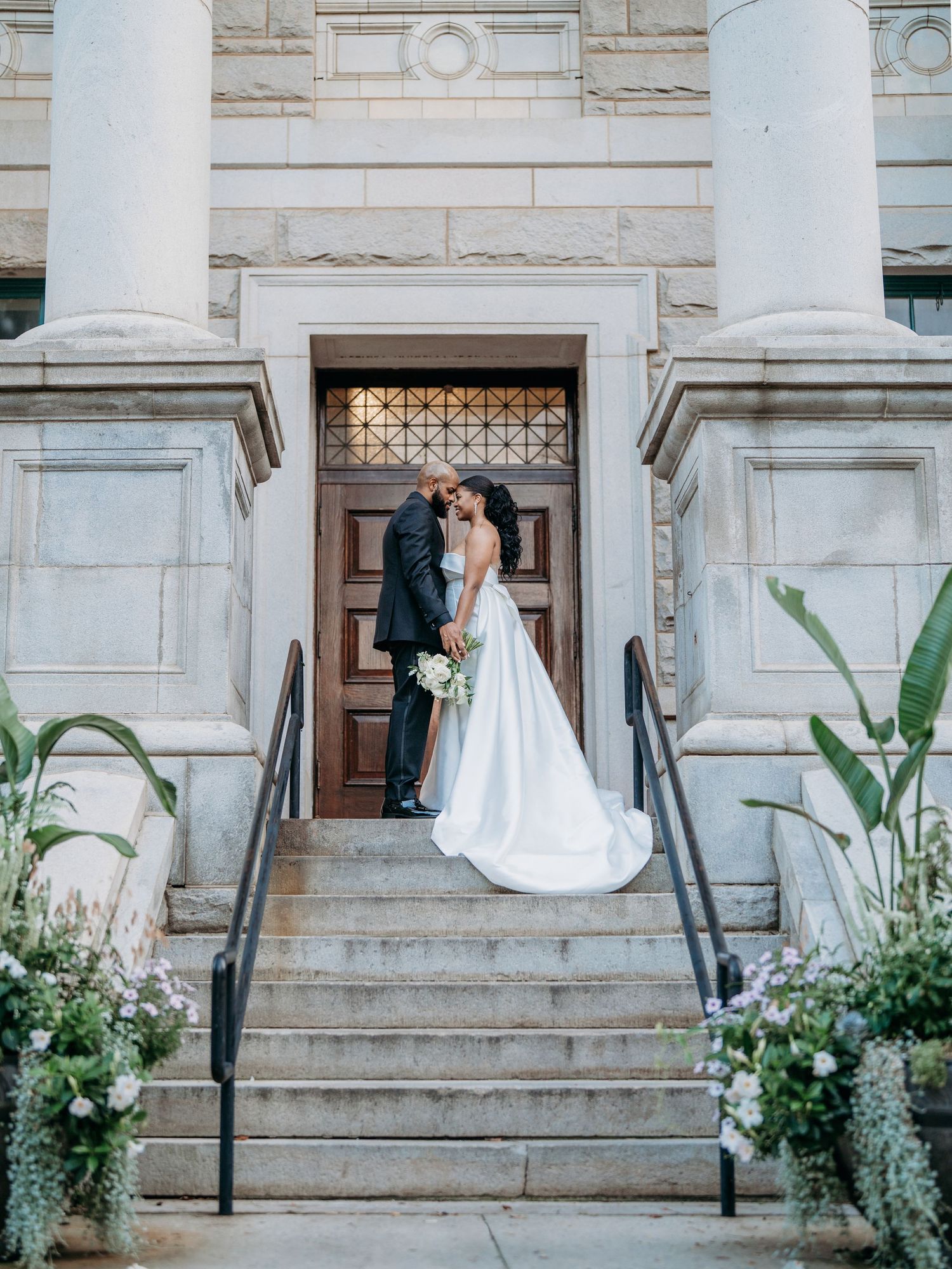 A bride in a flowing white gown stands elegantly on stone steps with white columns and greenery decorations.