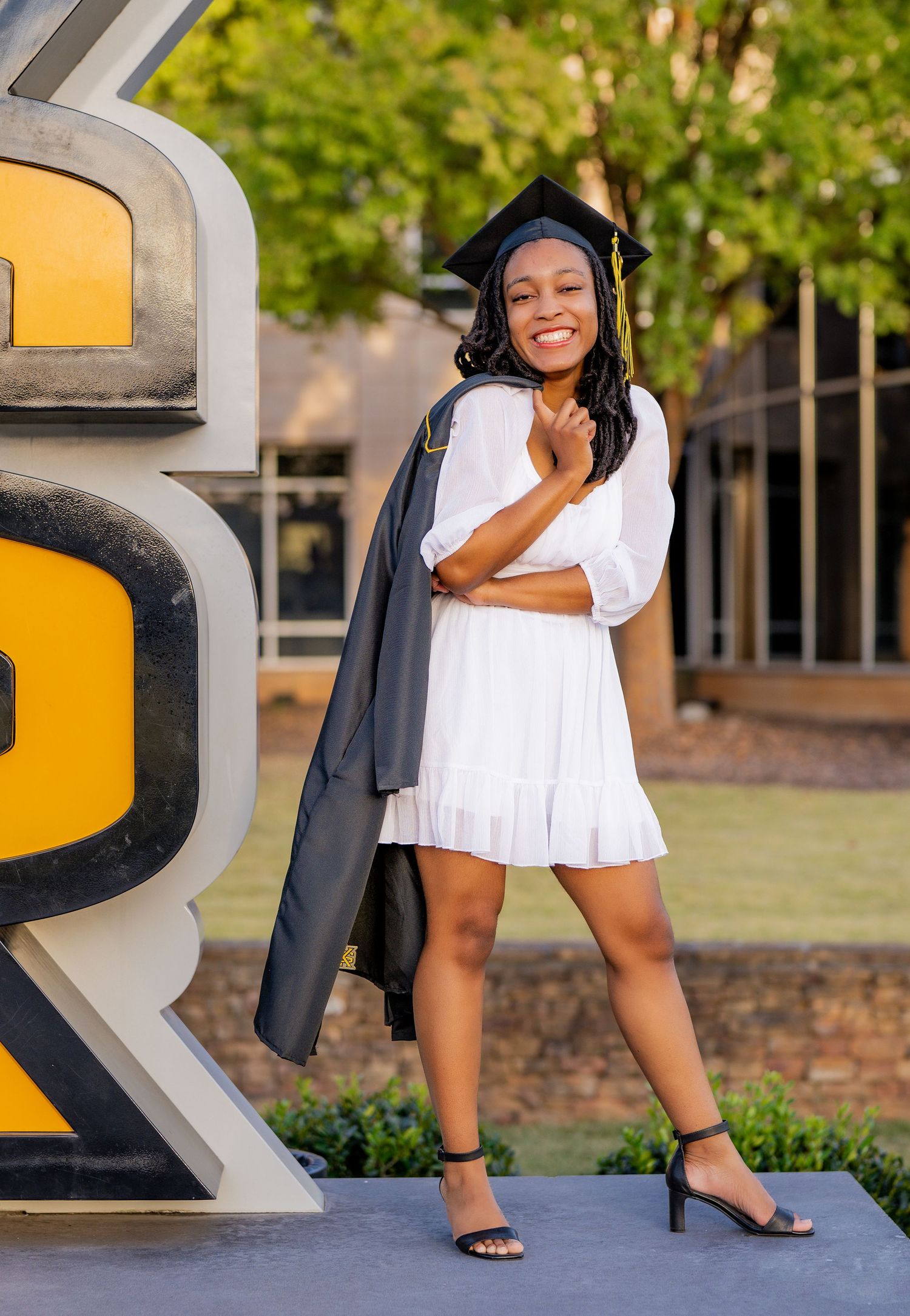 A person in a white dress and heels poses stylishly near large decorative letters in an outdoor setting.