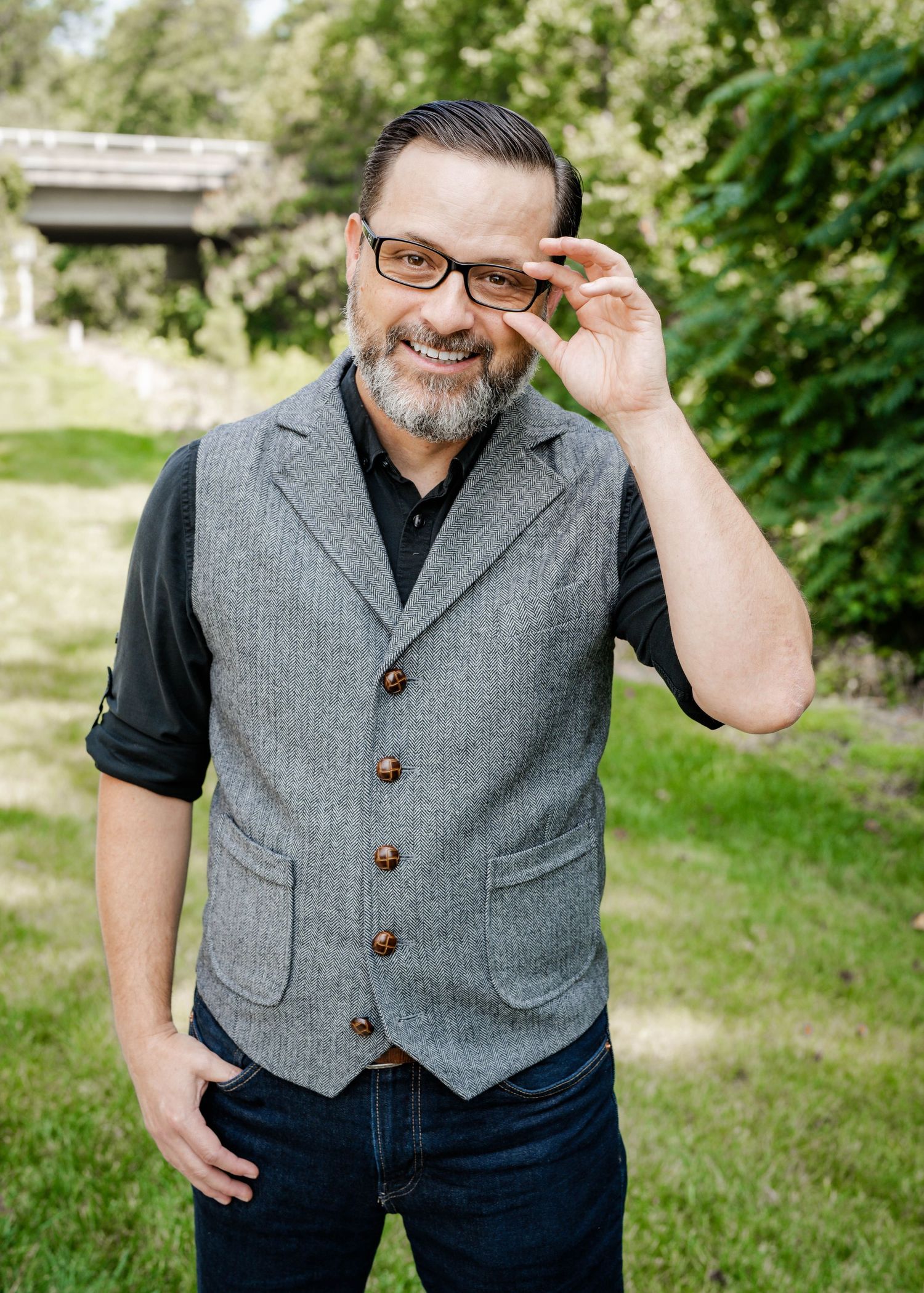 A person wearing glasses and a gray vest smiles while adjusting their frames in an outdoor garden setting.