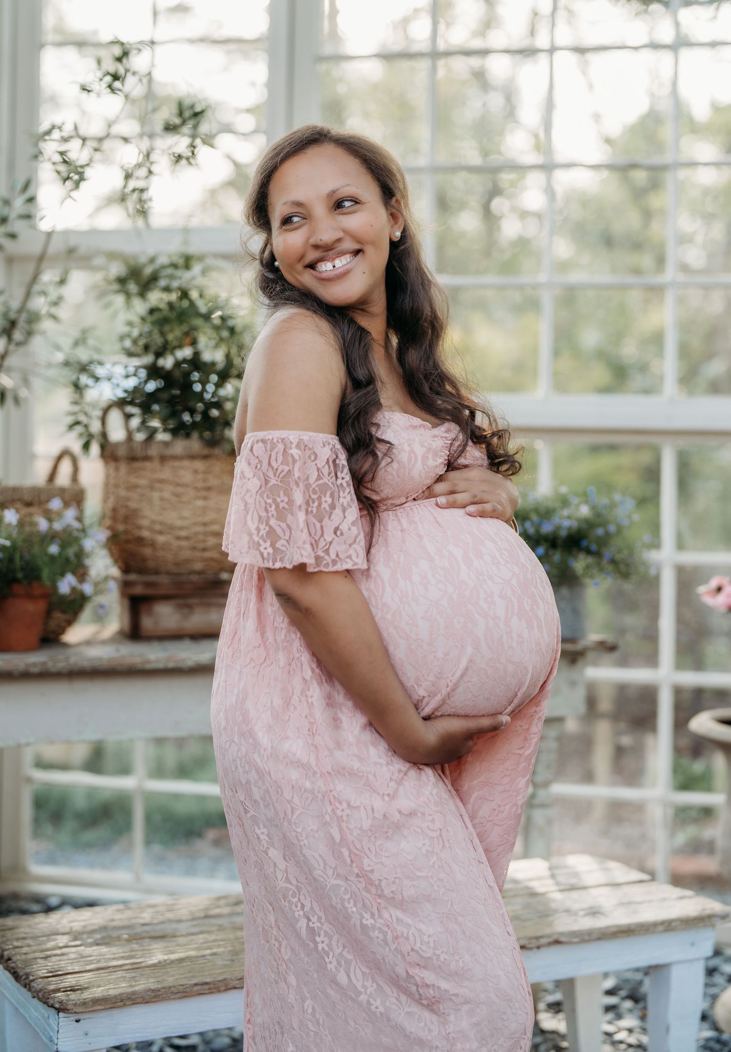 Expectant mother wearing pink lace off-shoulder maternity gown poses in sunlit greenhouse with plants and decor.