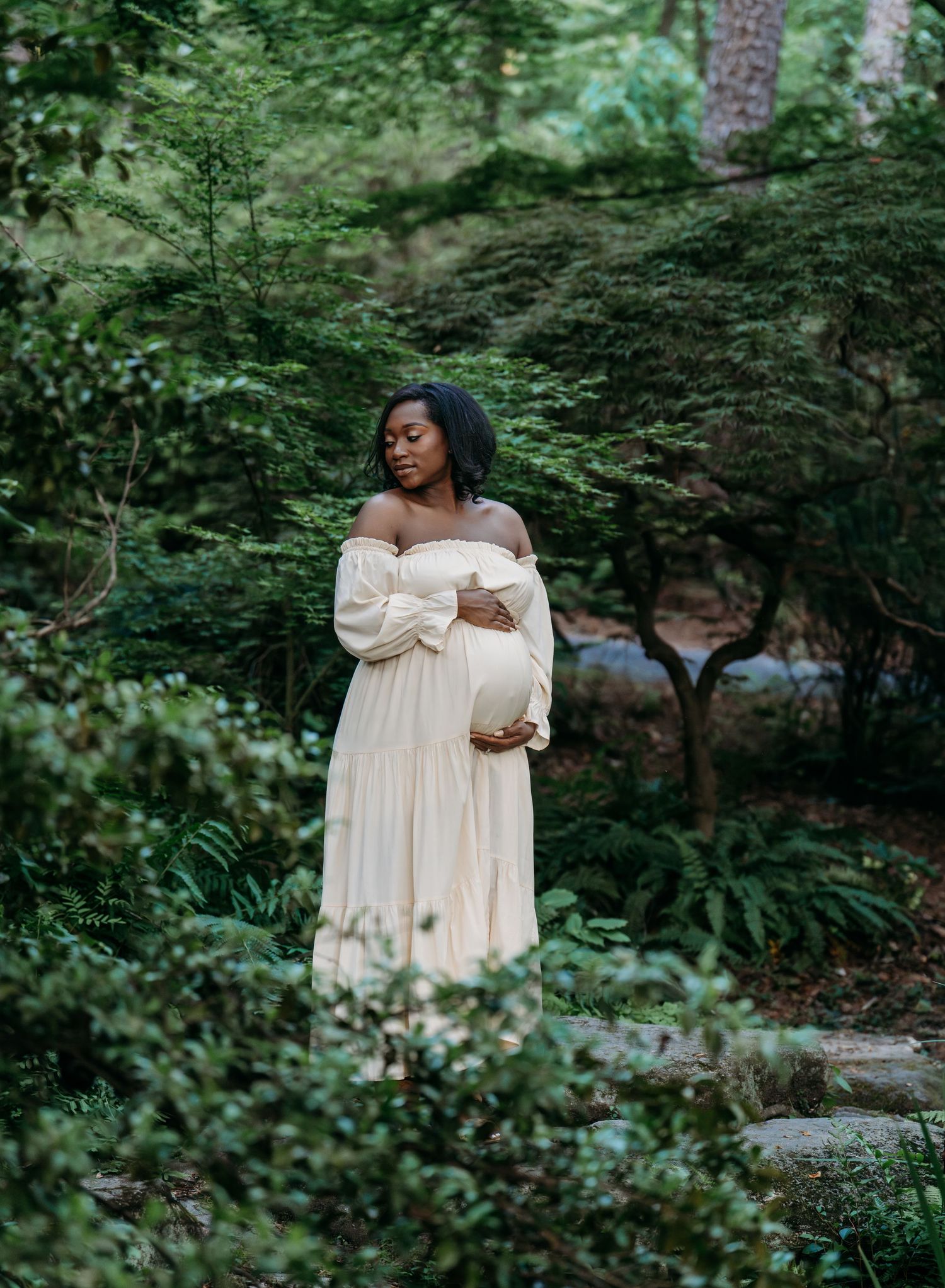 Expectant mother in flowing cream off-shoulder dress stands gracefully amid lush garden greenery and stone pathway.