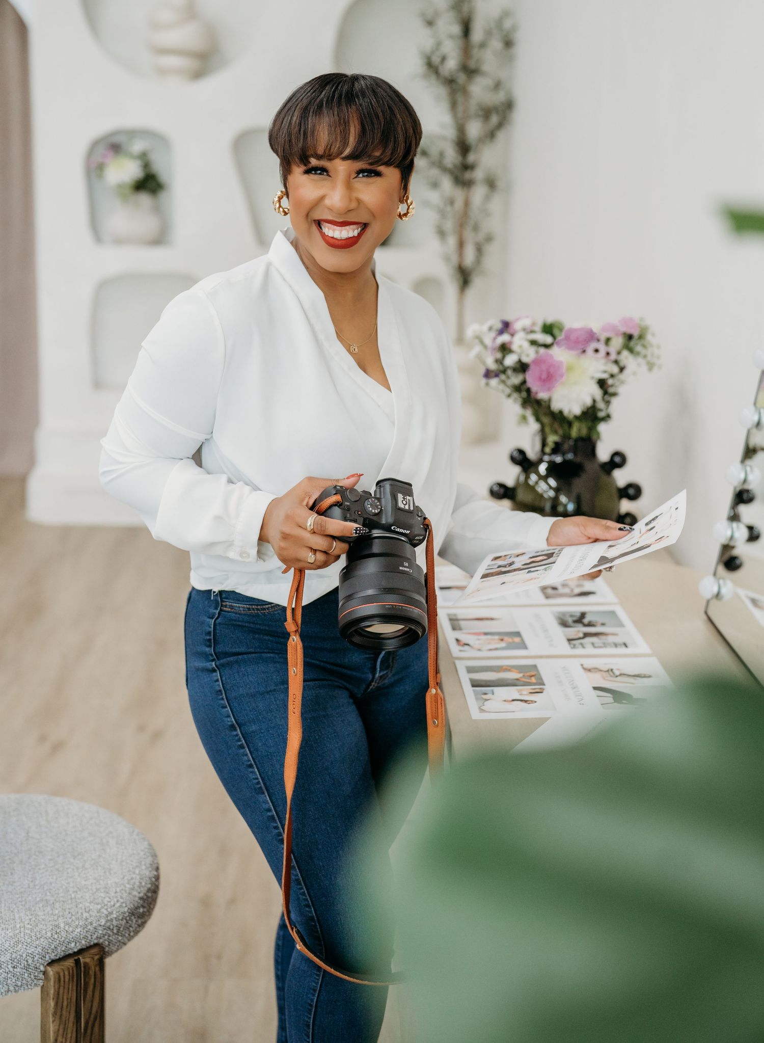 Professional photographer in white shirt and jeans holds DSLR camera while working in bright studio with floral decor.