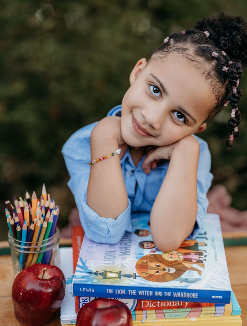 A young girl with braids smiles at a table with colored pencils, apples, and books including a Dictionary.