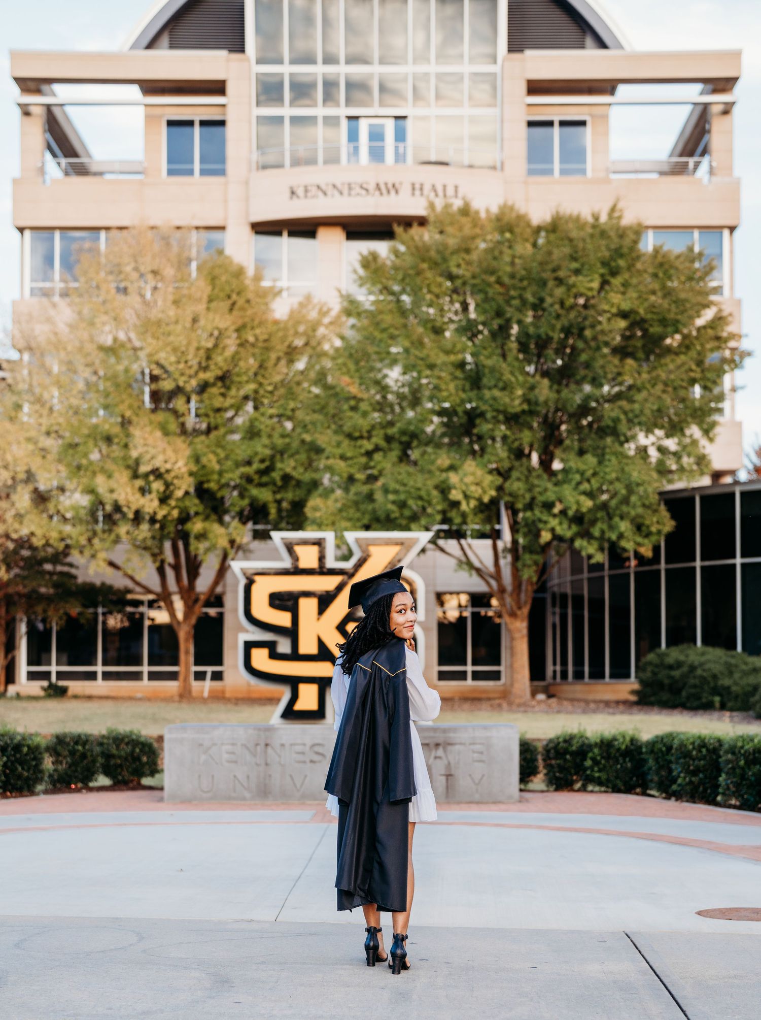 Graduate stands in front of Kennesaw Hall in dark green academic regalia during commencement photo session.