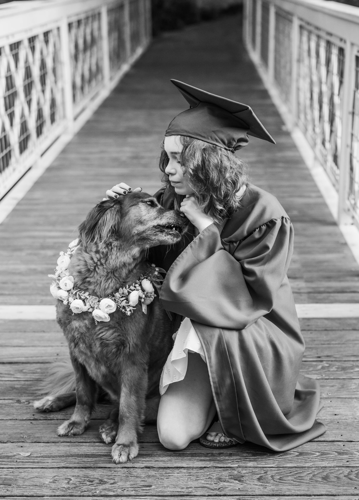 A graduate in cap and gown kneels on a wooden bridge sharing a tender moment with their dog wearing a floral wreath.