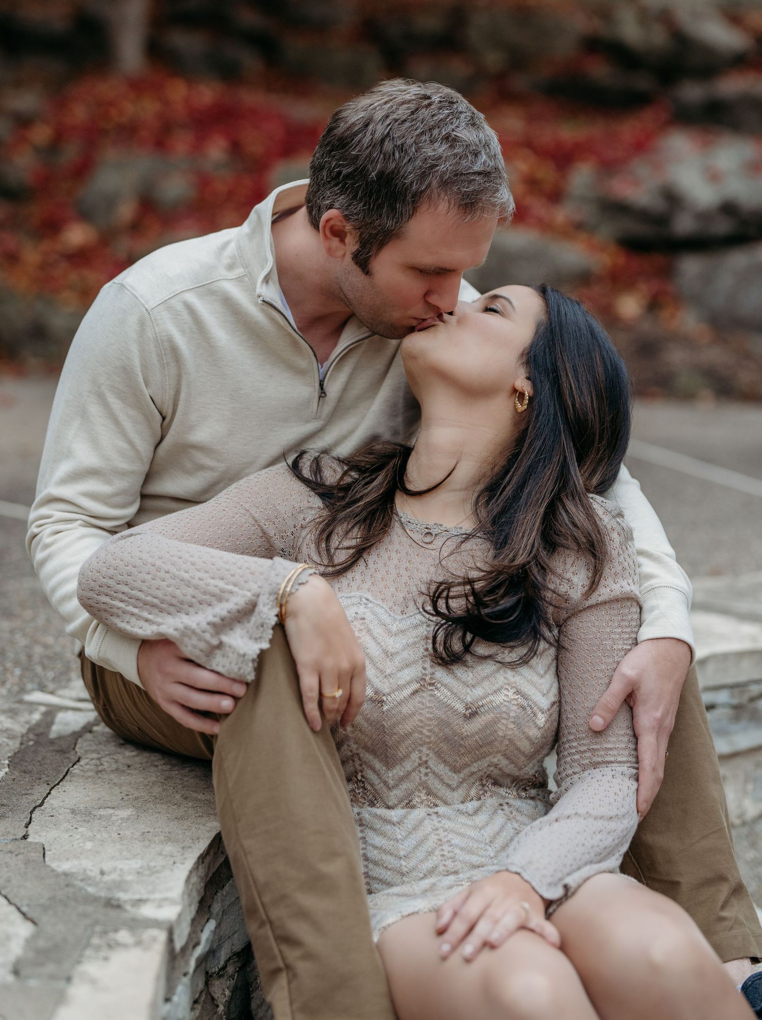 Couple in neutral outfits sharing an intimate kiss while sitting on stone steps surrounded by autumn foliage.