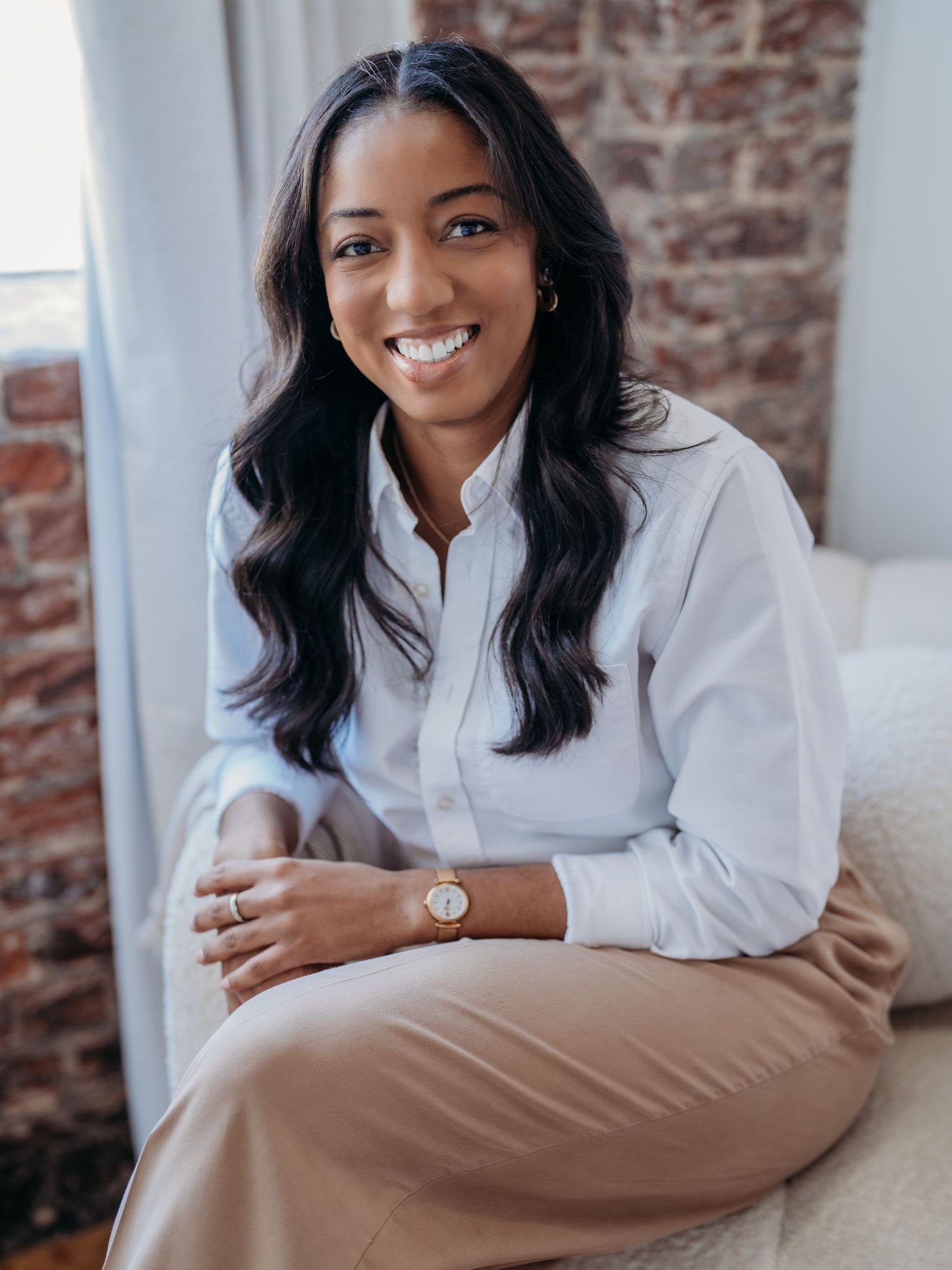 Professional portrait of someone in a white button-down shirt and khaki pants seated by an exposed brick wall.