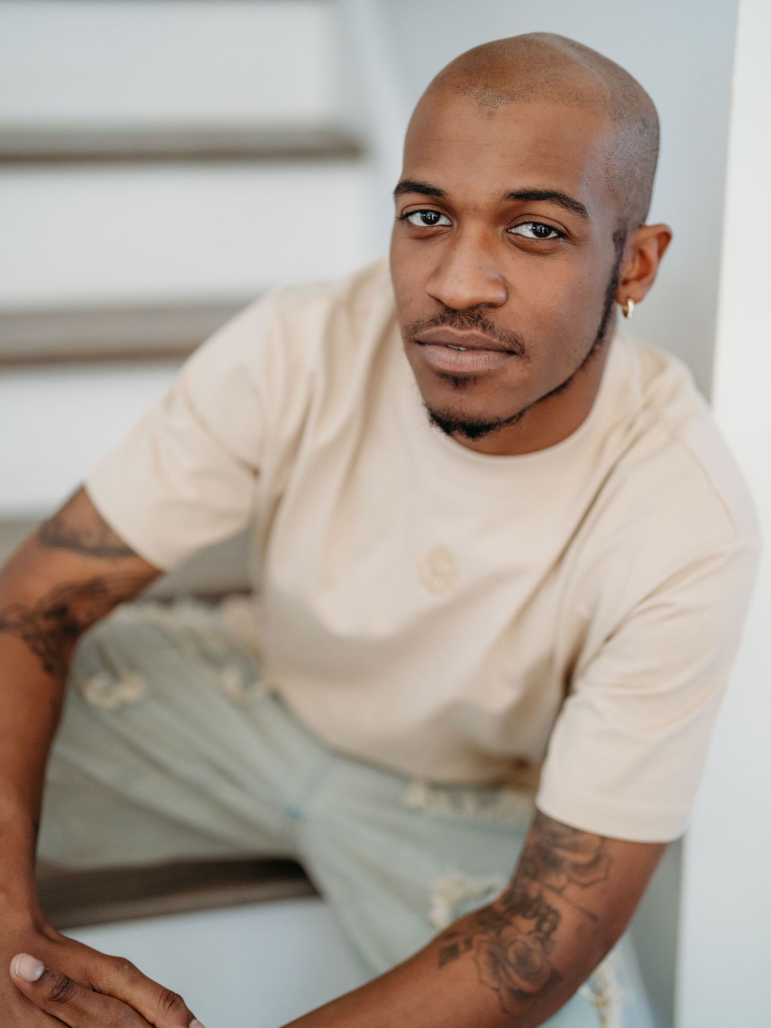A person in a beige t-shirt sits casually indoors showing visible arm tattoos against a light background.