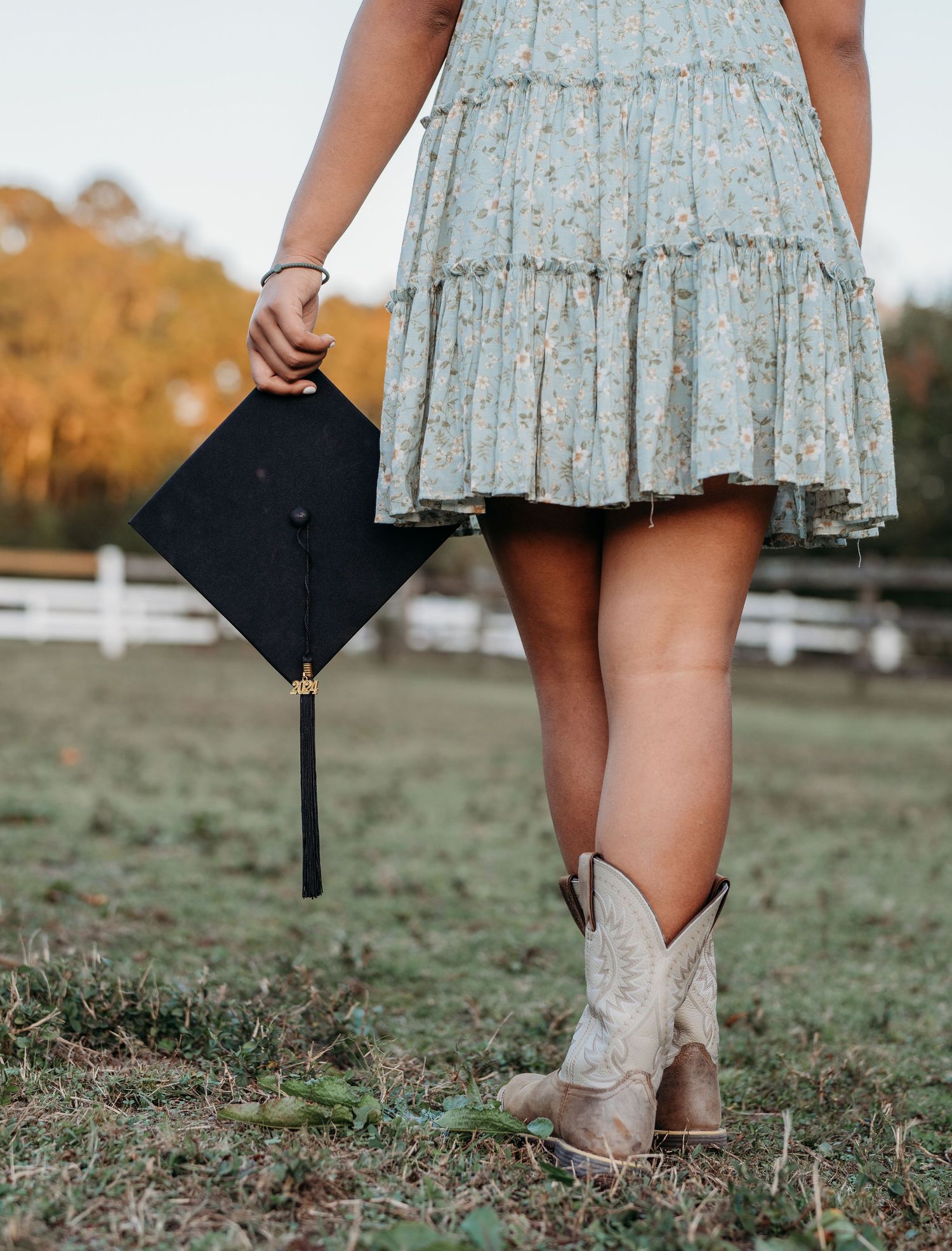 Graduate in floral dress and cowboy boots holds graduation cap in rustic outdoor setting with autumn trees and fence.