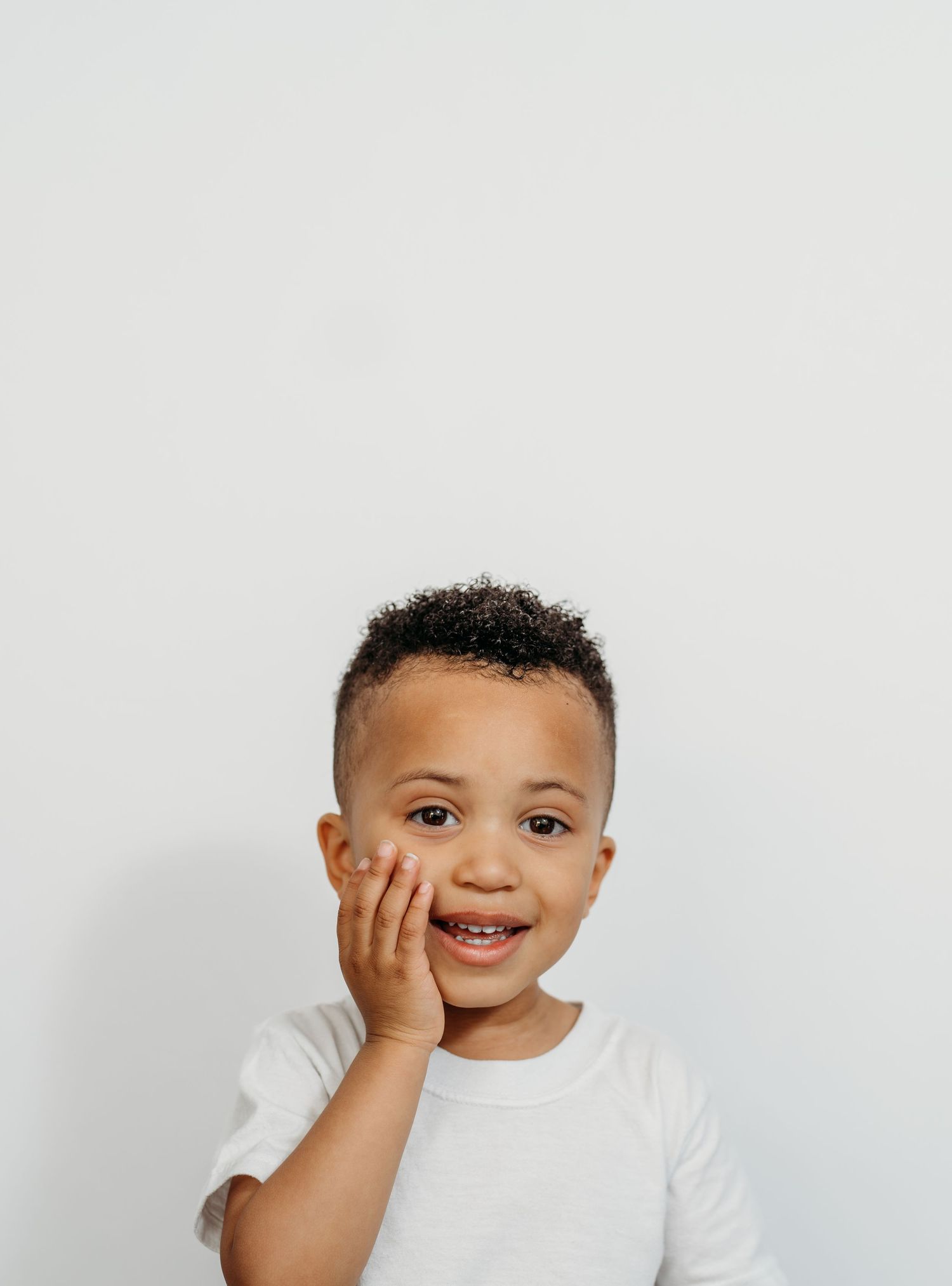 Young child in a white t-shirt smiles while touching their cheek against white background.