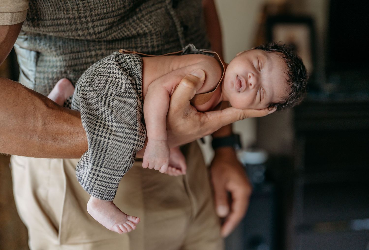 Person in a grey patterned shirt holds a tiny sleeping baby with pink feet and outfit.