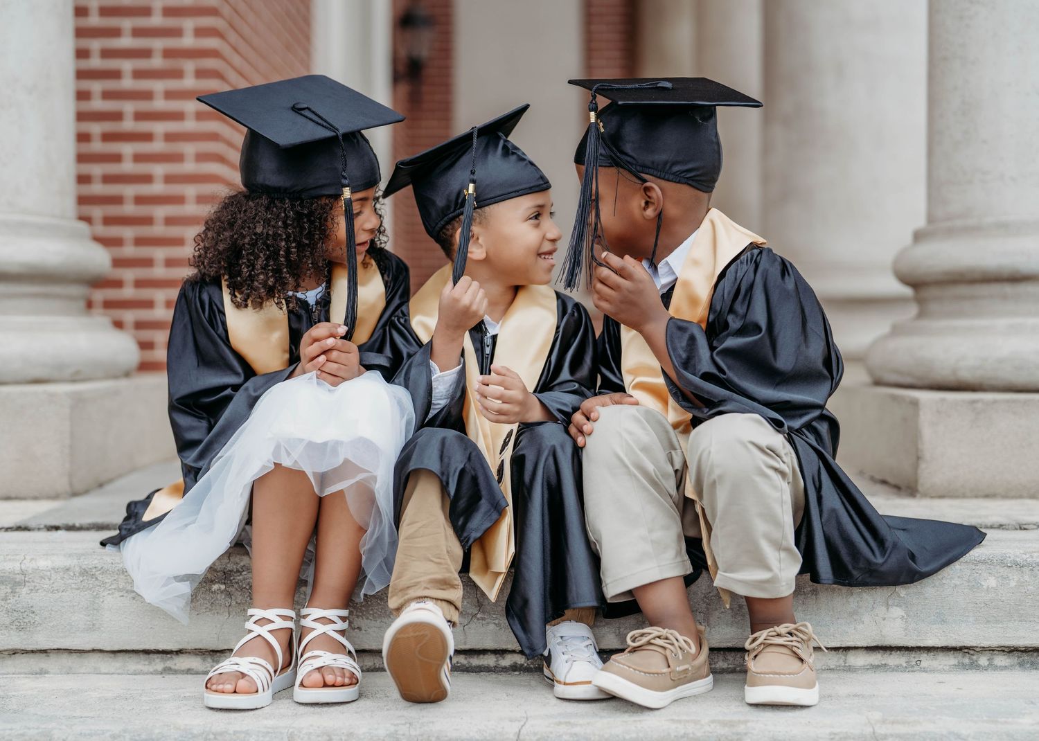 Three young children in graduation caps and gowns sit on steps, smiling and celebrating their academic achievement.