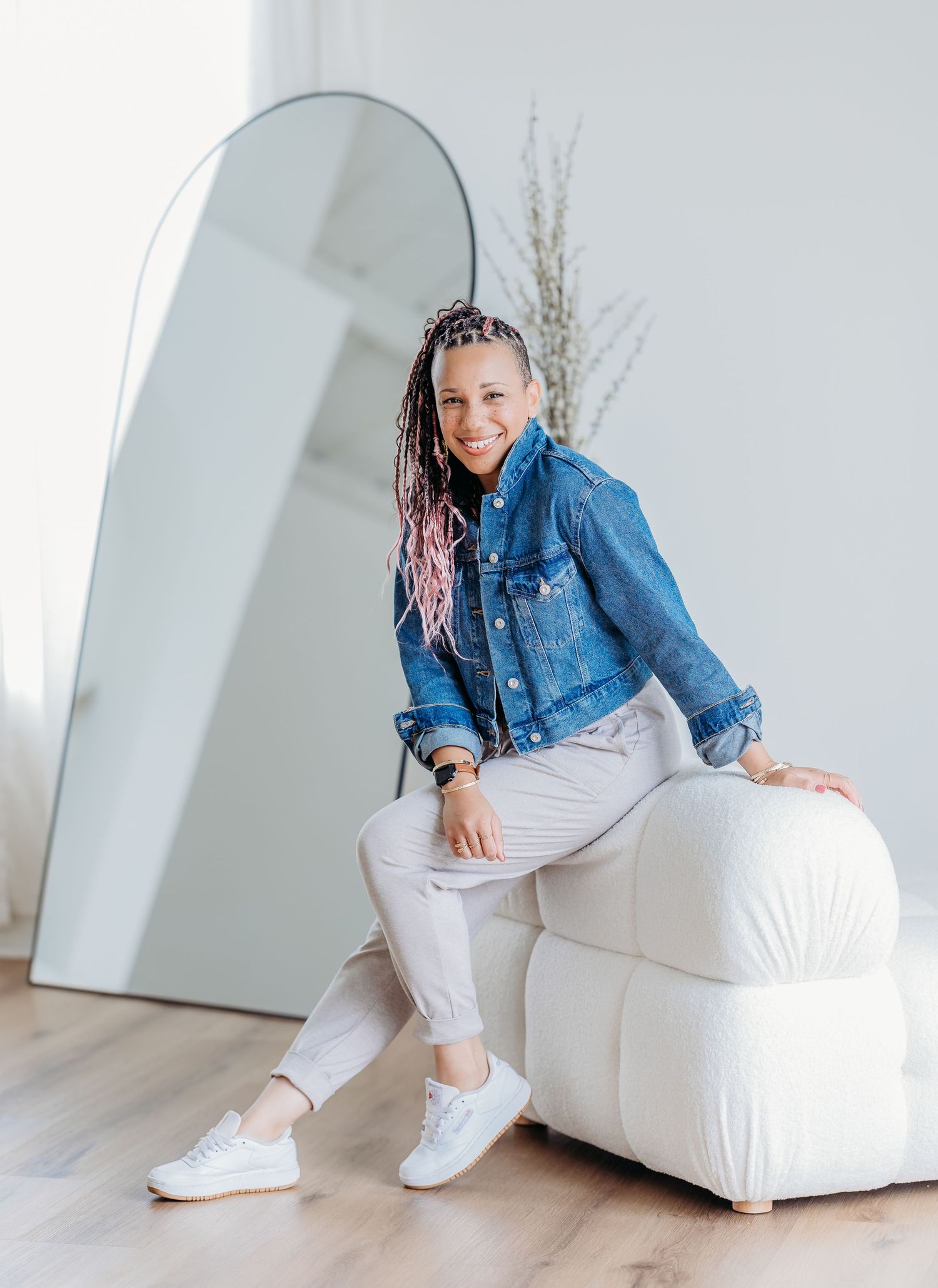 Casual lifestyle portrait of someone wearing a denim jacket and white pants sitting on a white ottoman in a bright room.