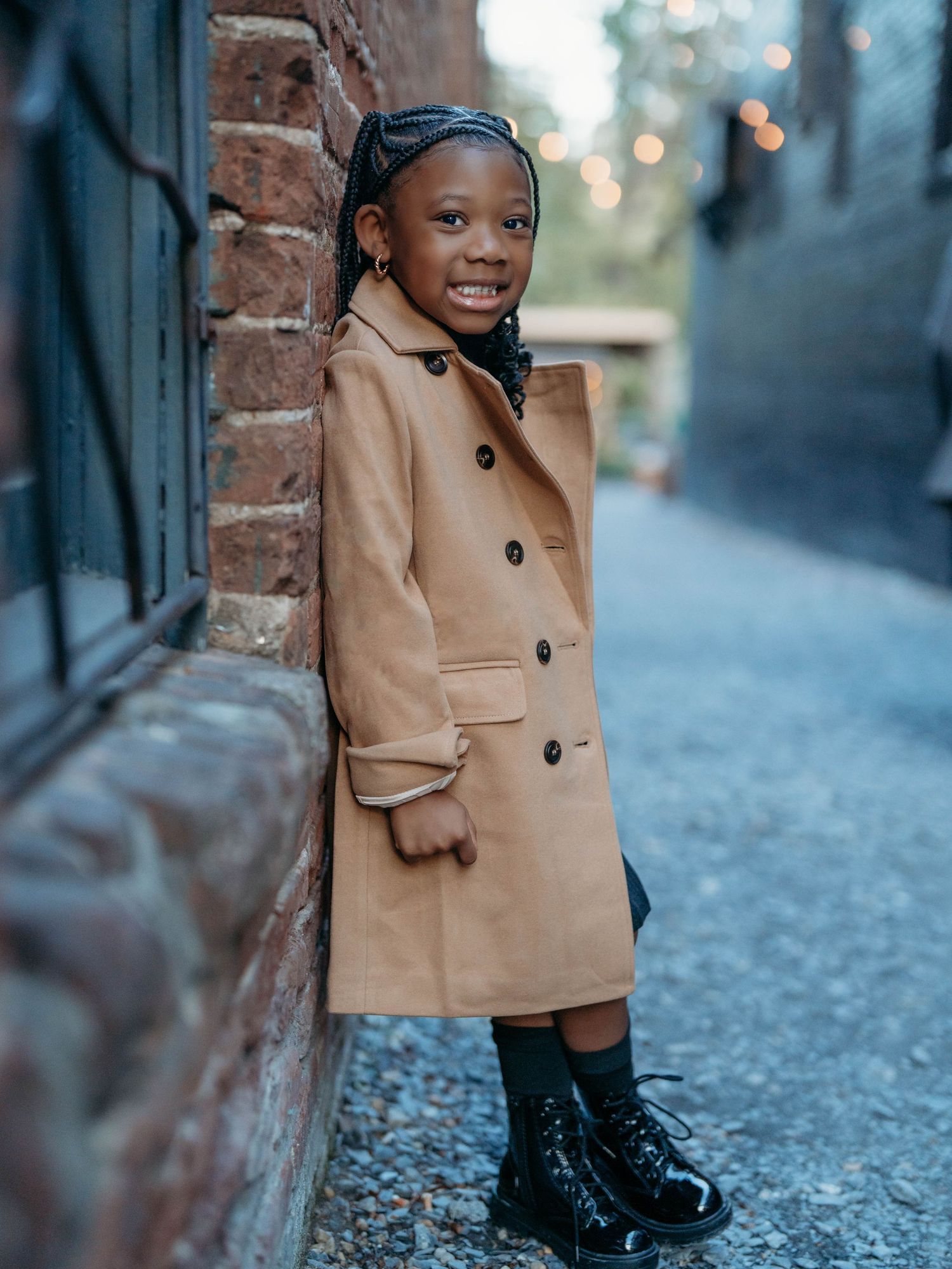 A child wearing a tan peacoat poses on a cobblestone street with brick buildings in the background.