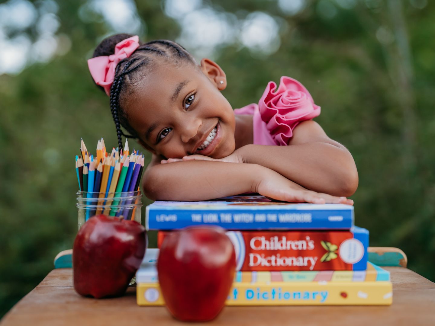 Smiling young girl resting on stack of childrens dictionary books with pencils and red apples on wooden desk outdoors.