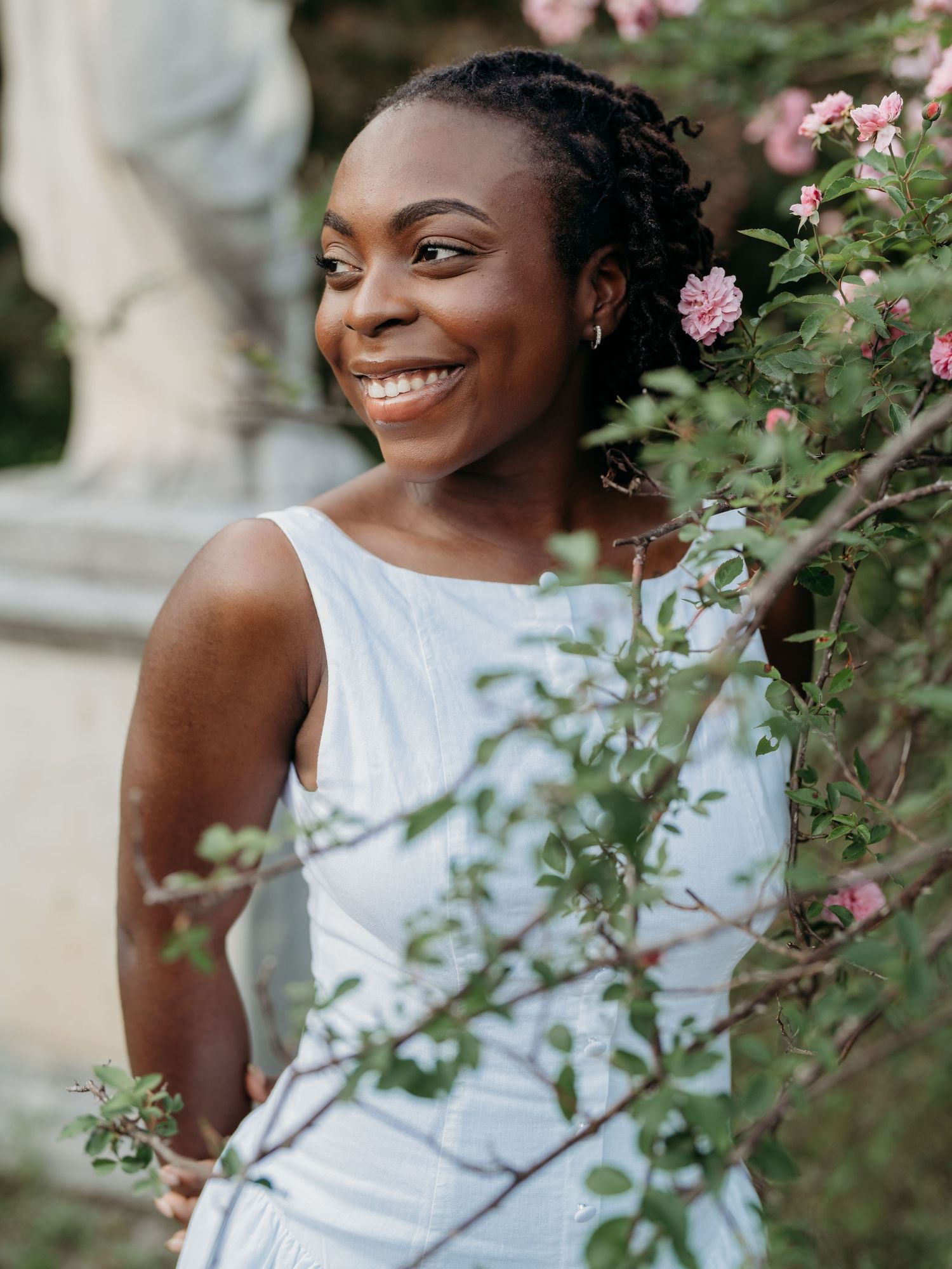 A joyful person in a white dress poses amid blooming pink roses in a garden setting.