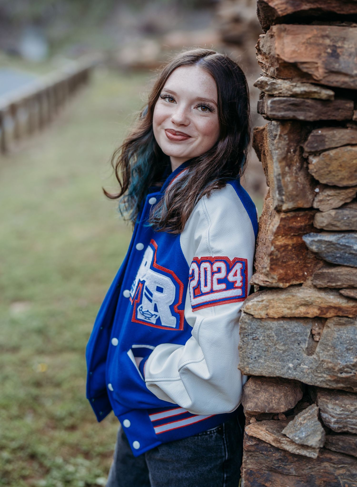 A varsity letterman jacket in blue and white features class of 2024 details against a rustic stone wall backdrop.
