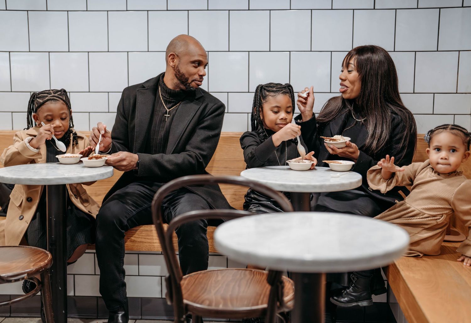 An African-American Family sit at small round tables in a restaurant with white subway tile walls.