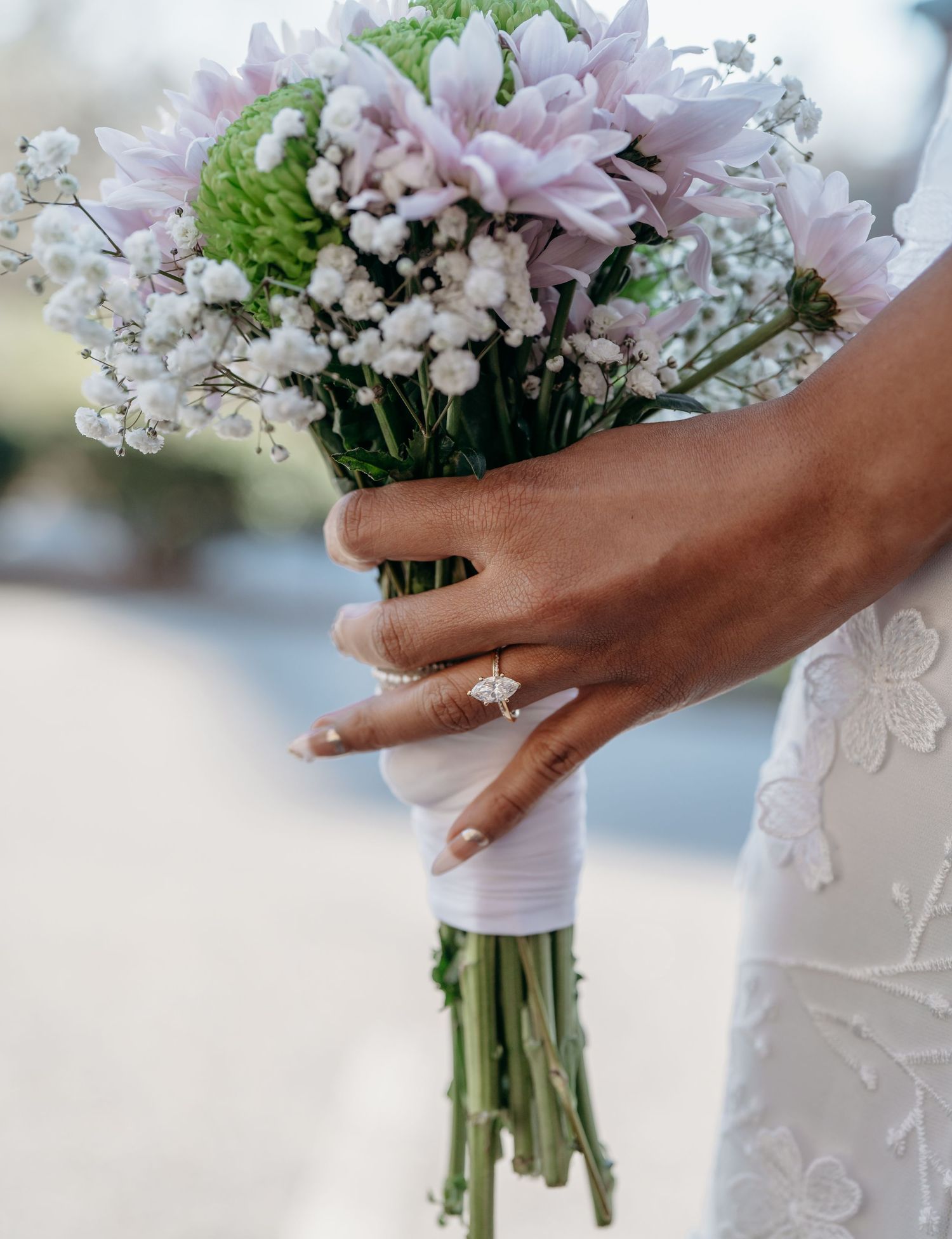 A hand wearing a glittering engagement ring holds a delicate bouquet of pink lilies, green buds and baby's breath flowers.